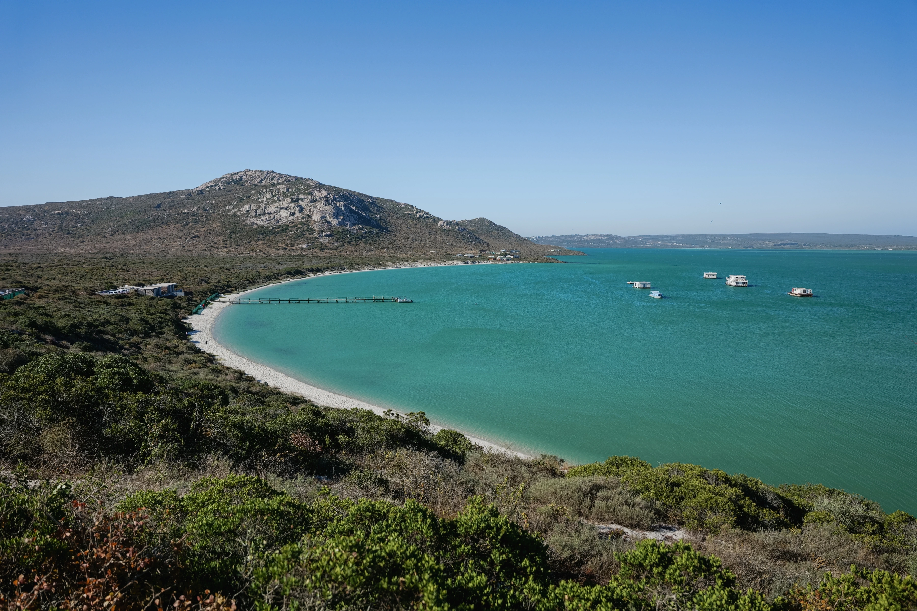 Small white boats and a pier sit in a turquoise bay bordered by pale shore, scrub and a rocky hill.