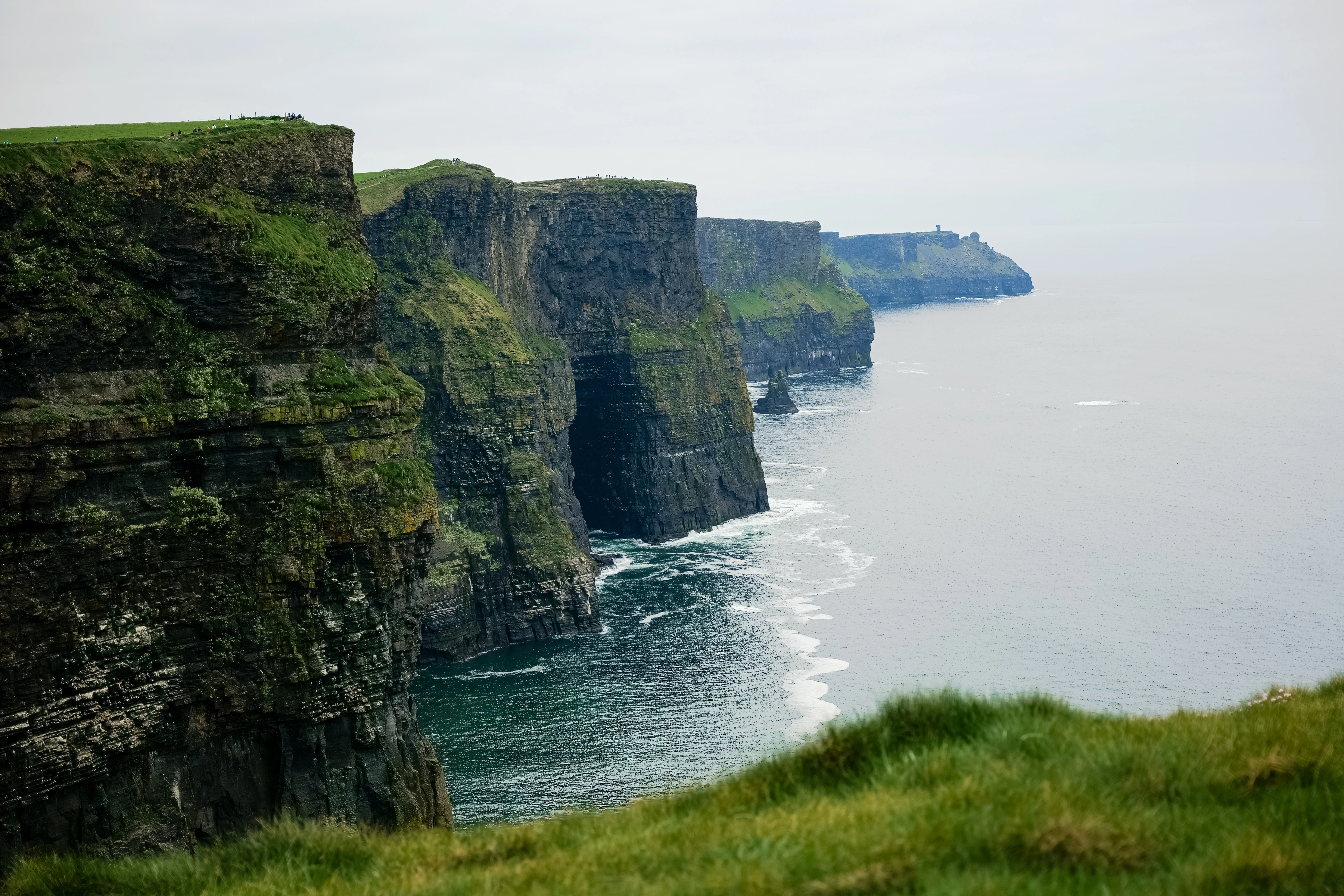 Layered sea cliffs recede into haze above blue water, with small figures on the ridge.
