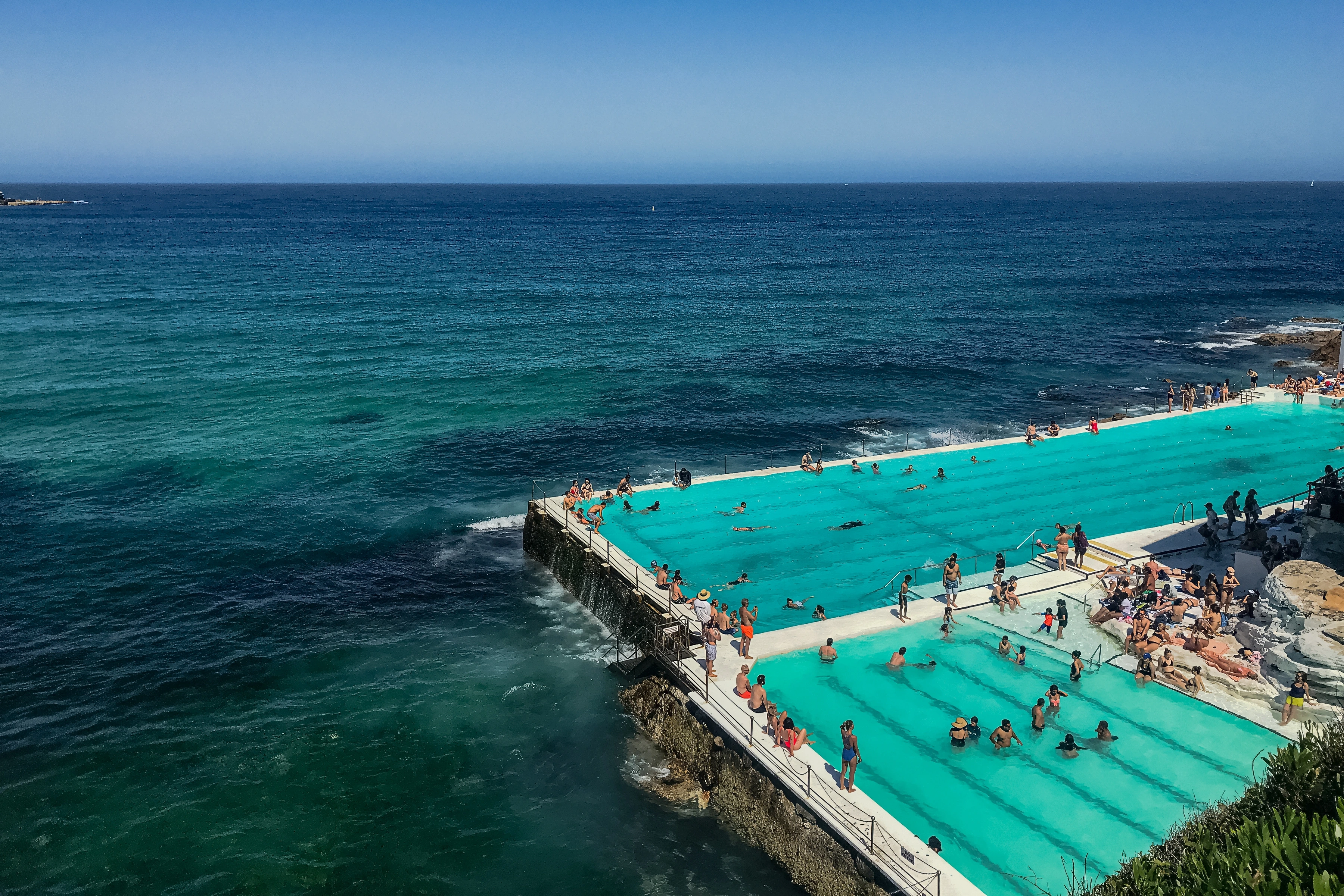 Bathers occupy a turquoise tidal pool beside darker open sea.