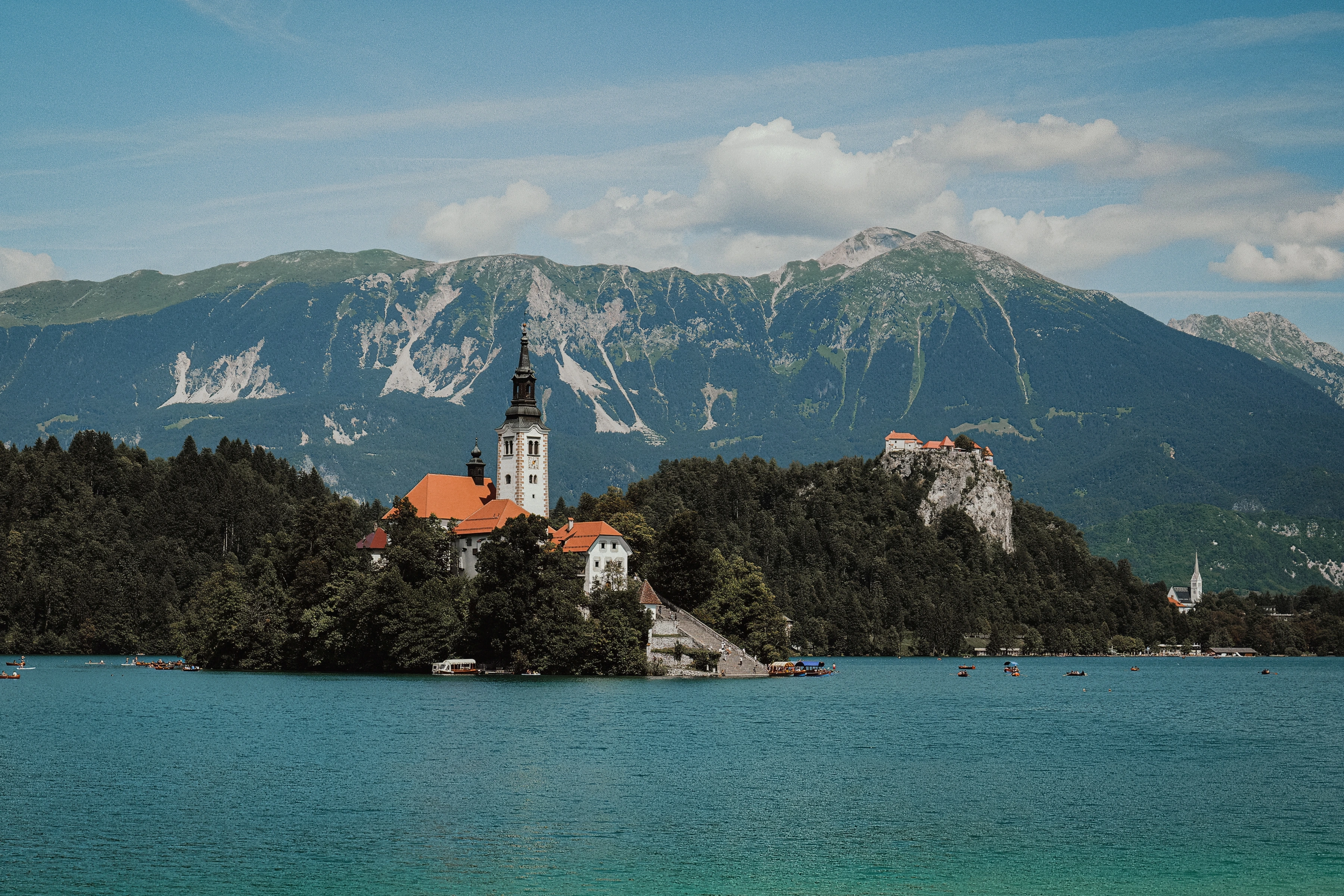 A church and red-roofed buildings occupy a wooded island, with a cliff-top building and mountains across the lake.