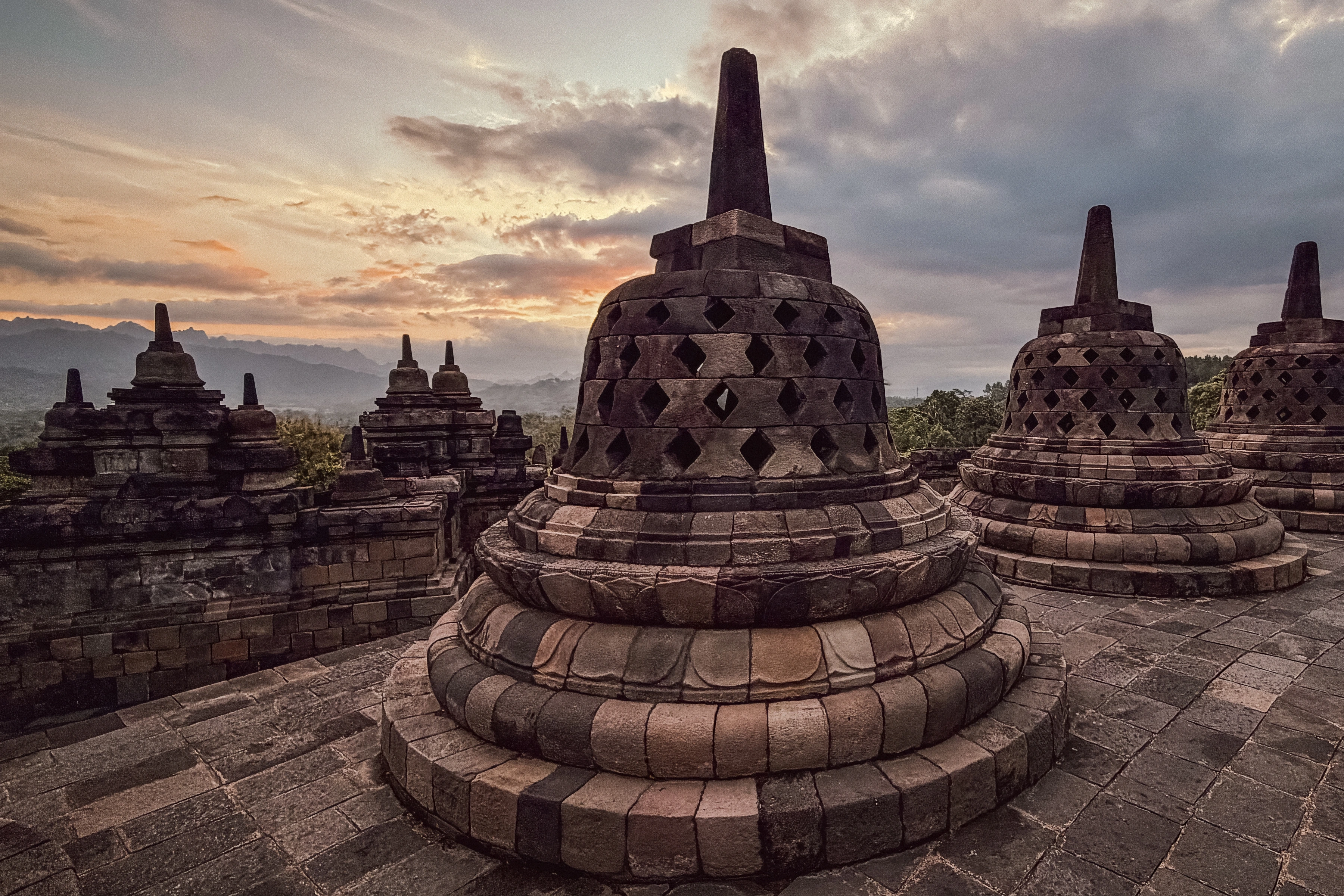 Perforated stone stupas repeat across a terrace beneath a warm opening in the clouds.