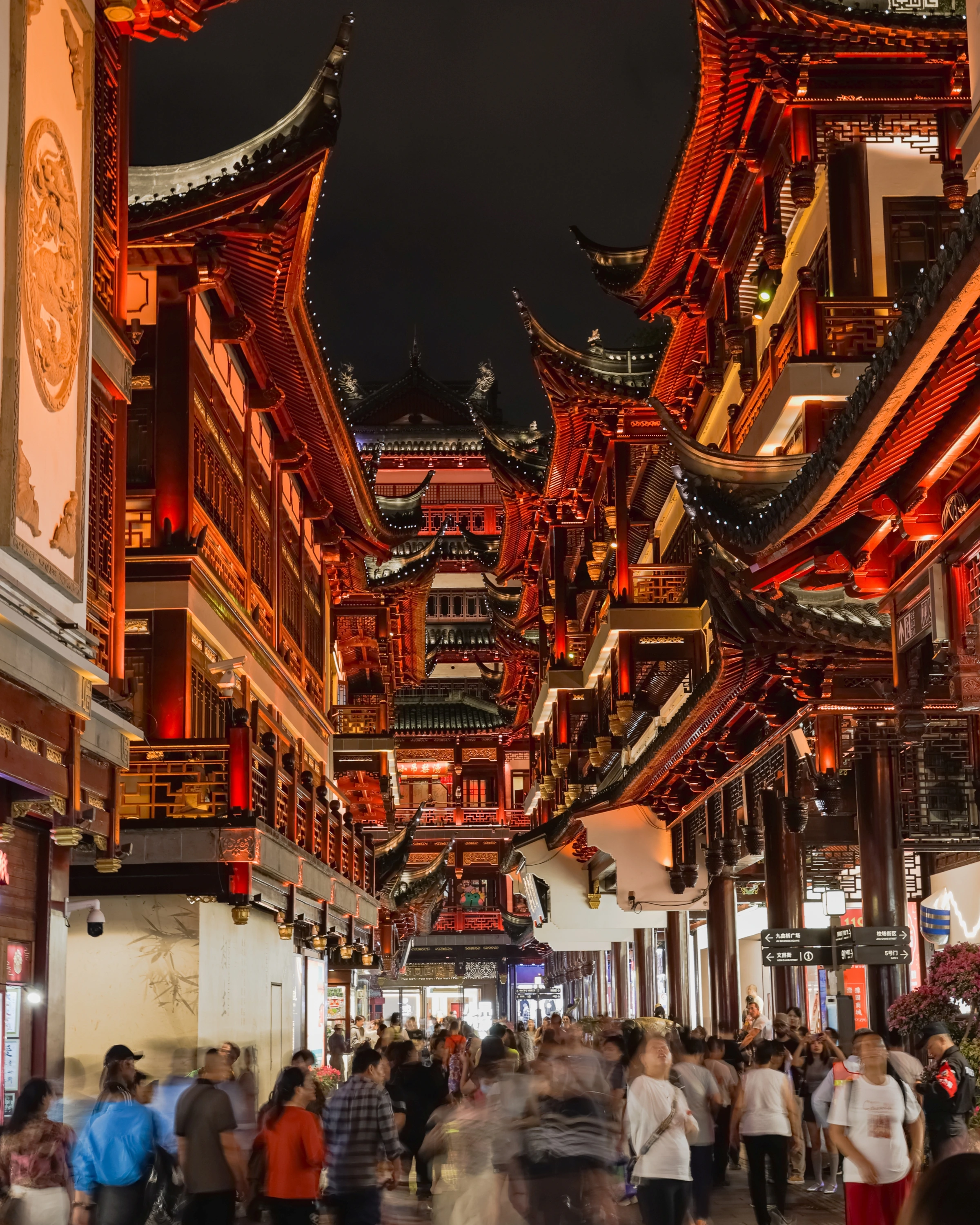Pedestrians pass beneath illuminated red and orange rooflines at night.