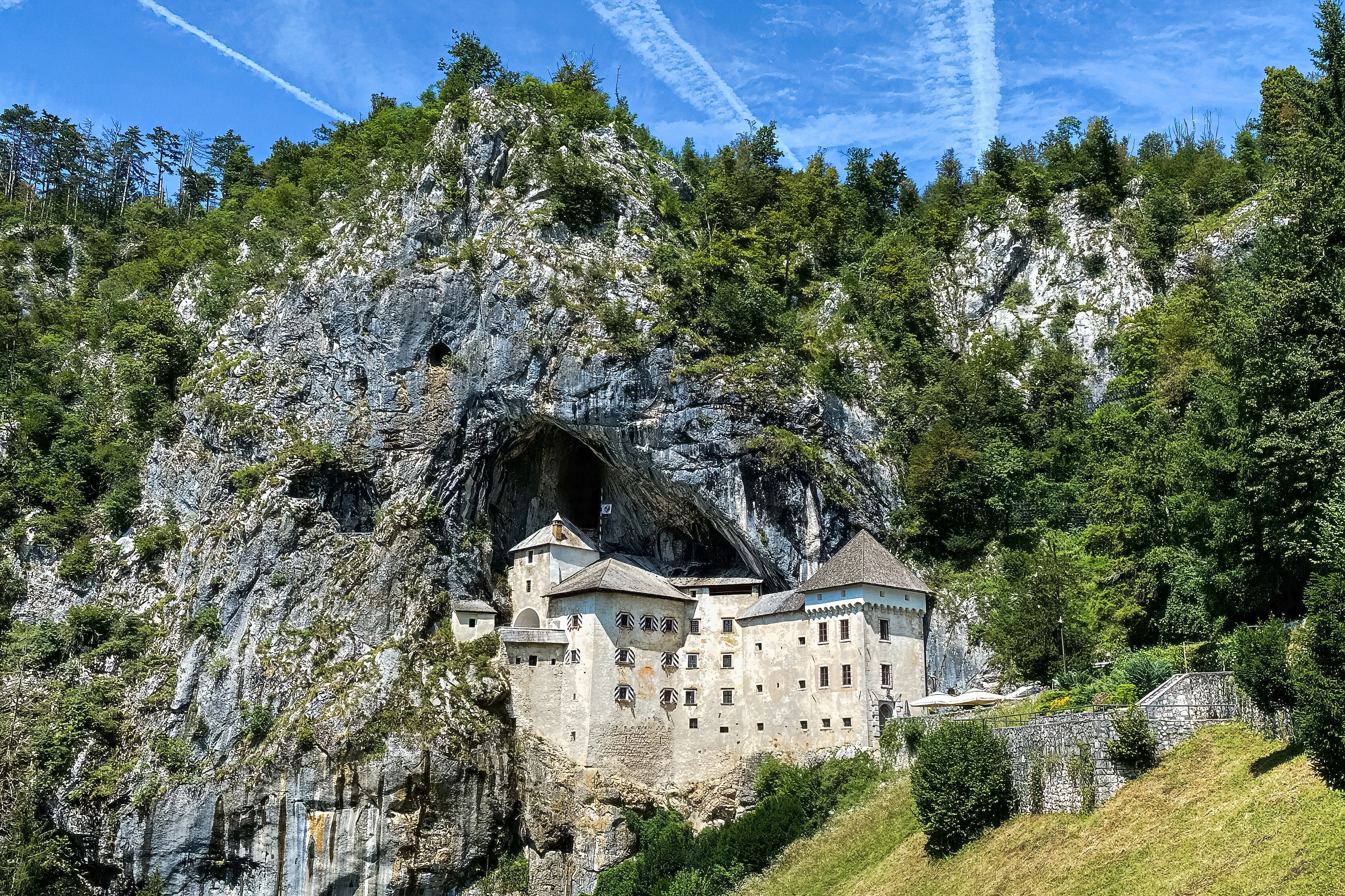 A pale castle occupies a dark recess in a sheer rocky cliff above green vegetation.