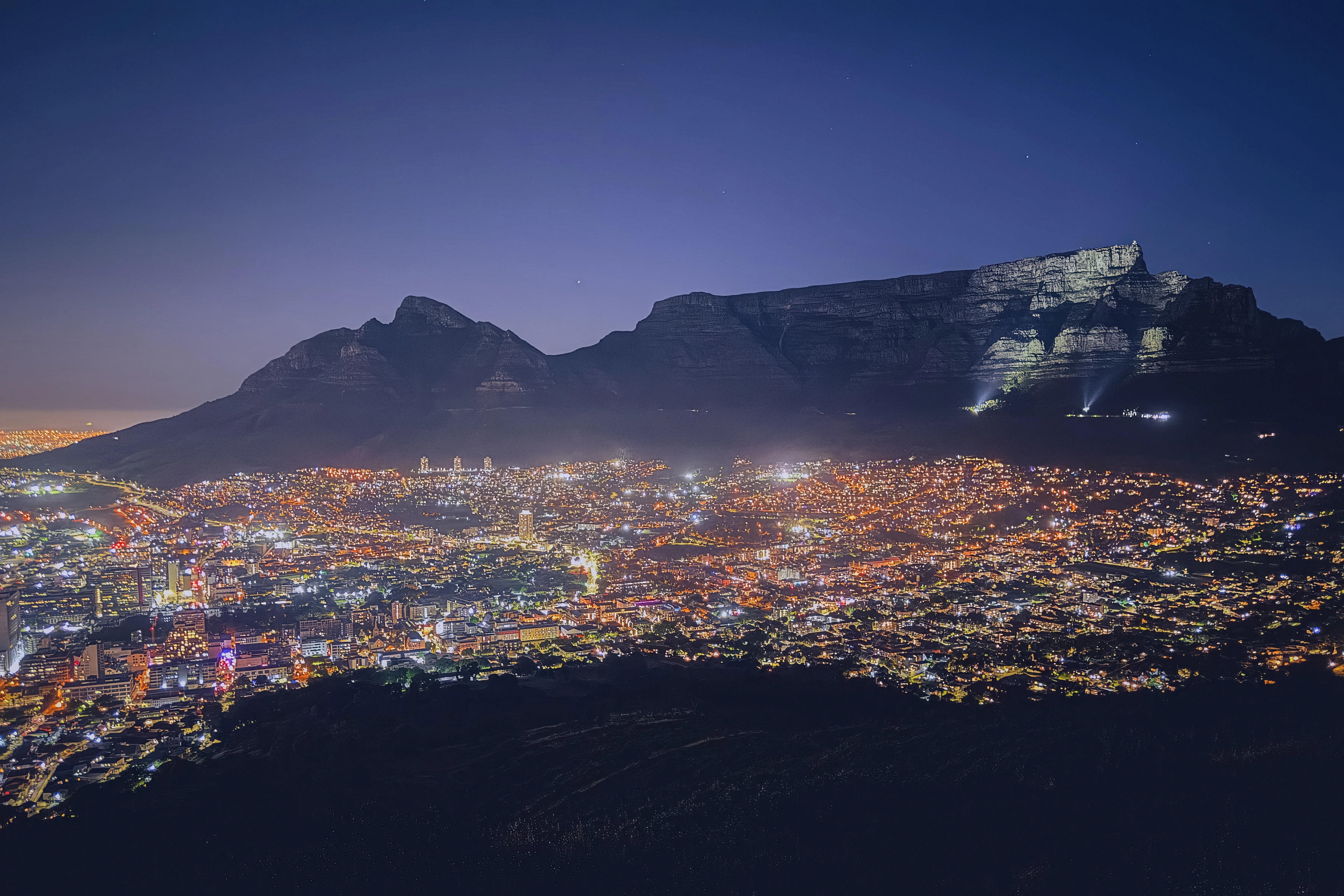 A network of city lights spreads below the dark mountain profile and a deep blue sky.