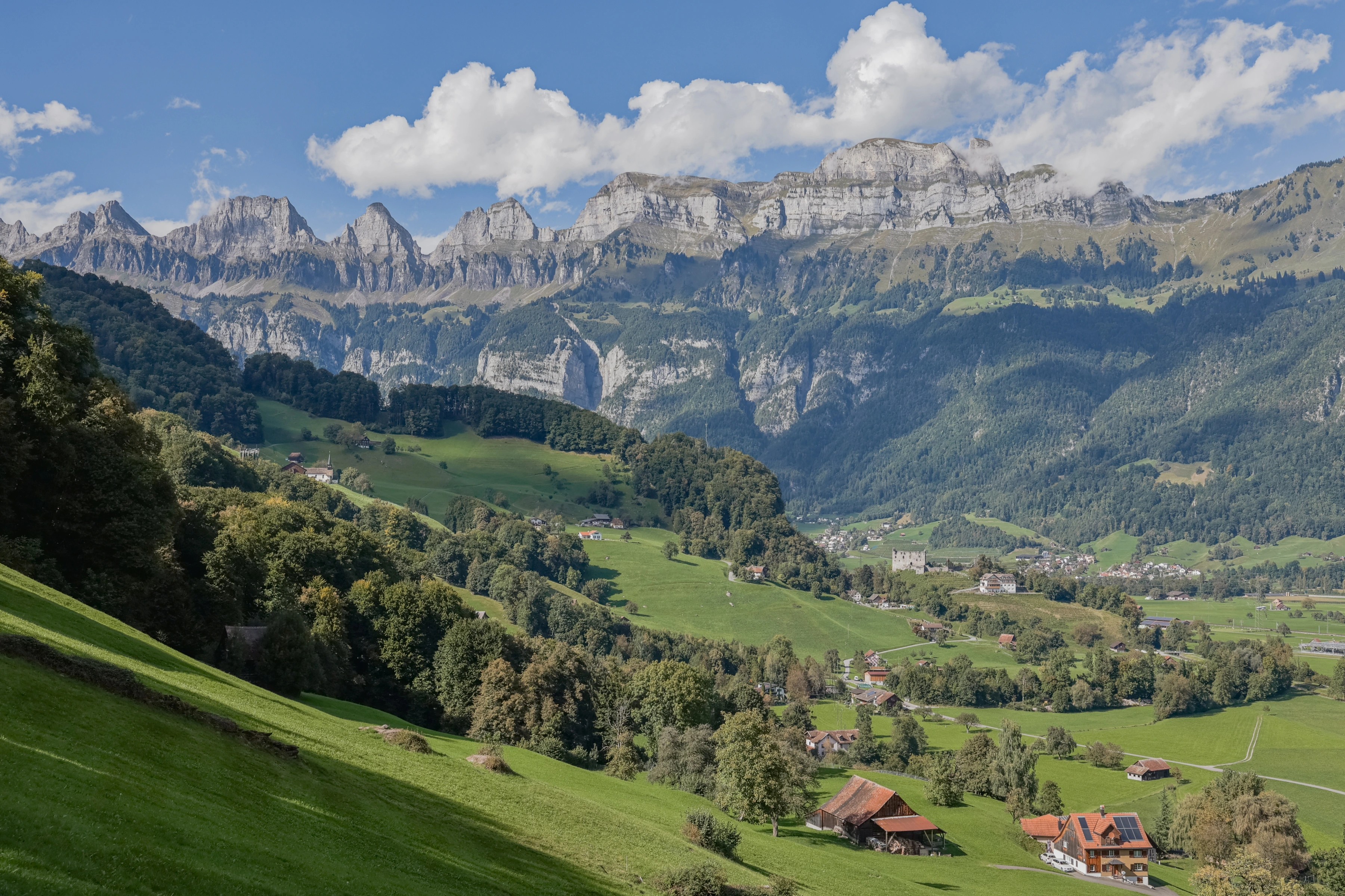 Scattered farm buildings and a larger stone structure sit among green pasture and forest beneath a craggy mountain wall.
