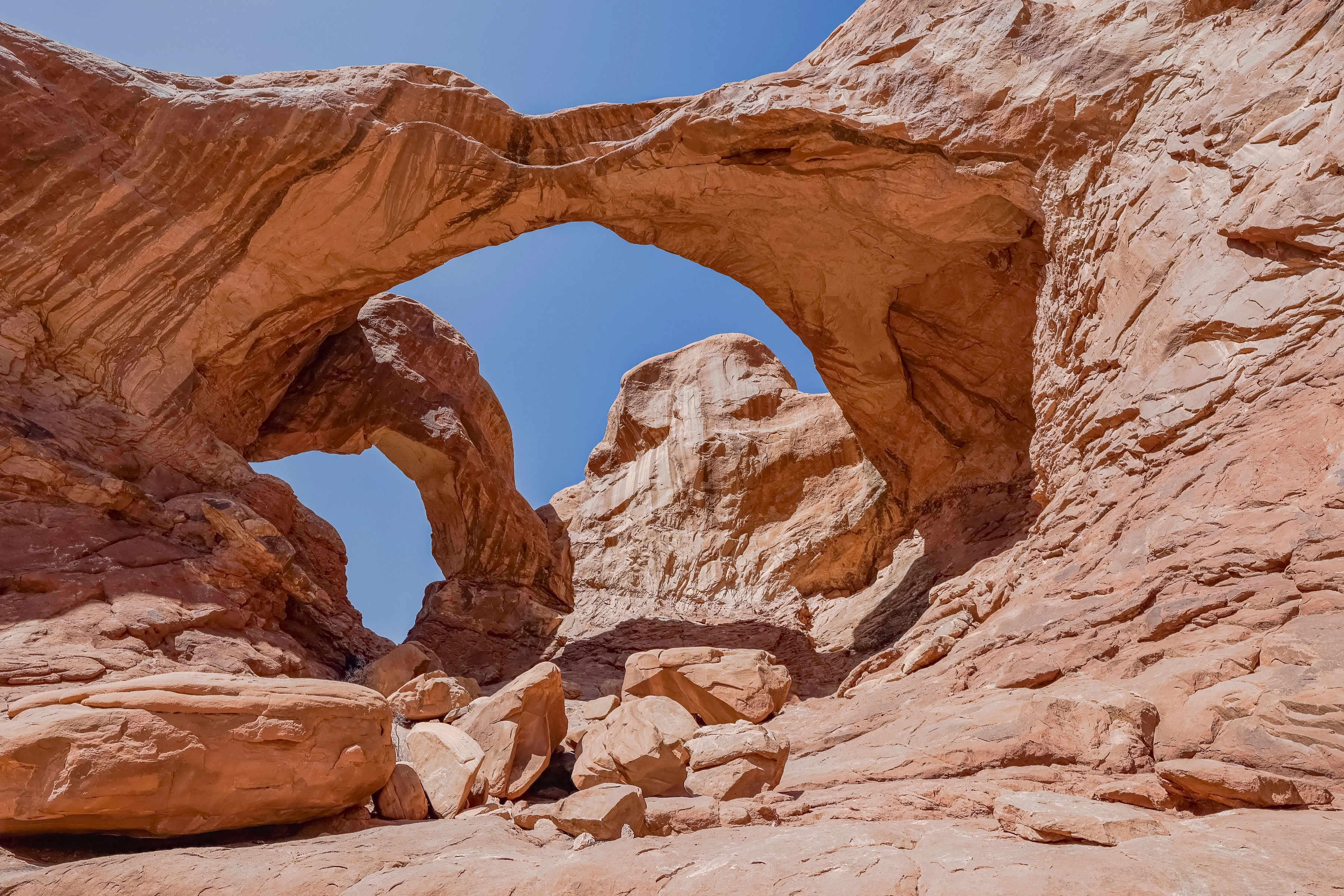 Warm orange rock forms two large arch openings against blue sky, with boulders below.