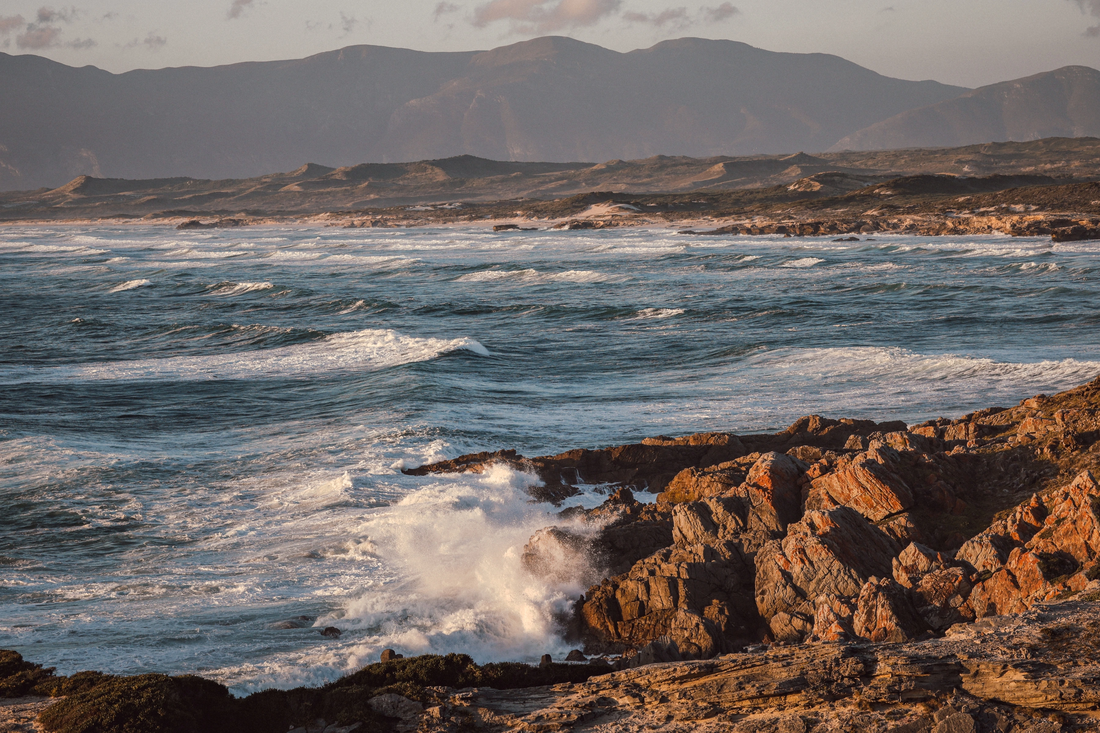 White water strikes warm orange coastal rock, with successive blue waves and a distant ridge behind.