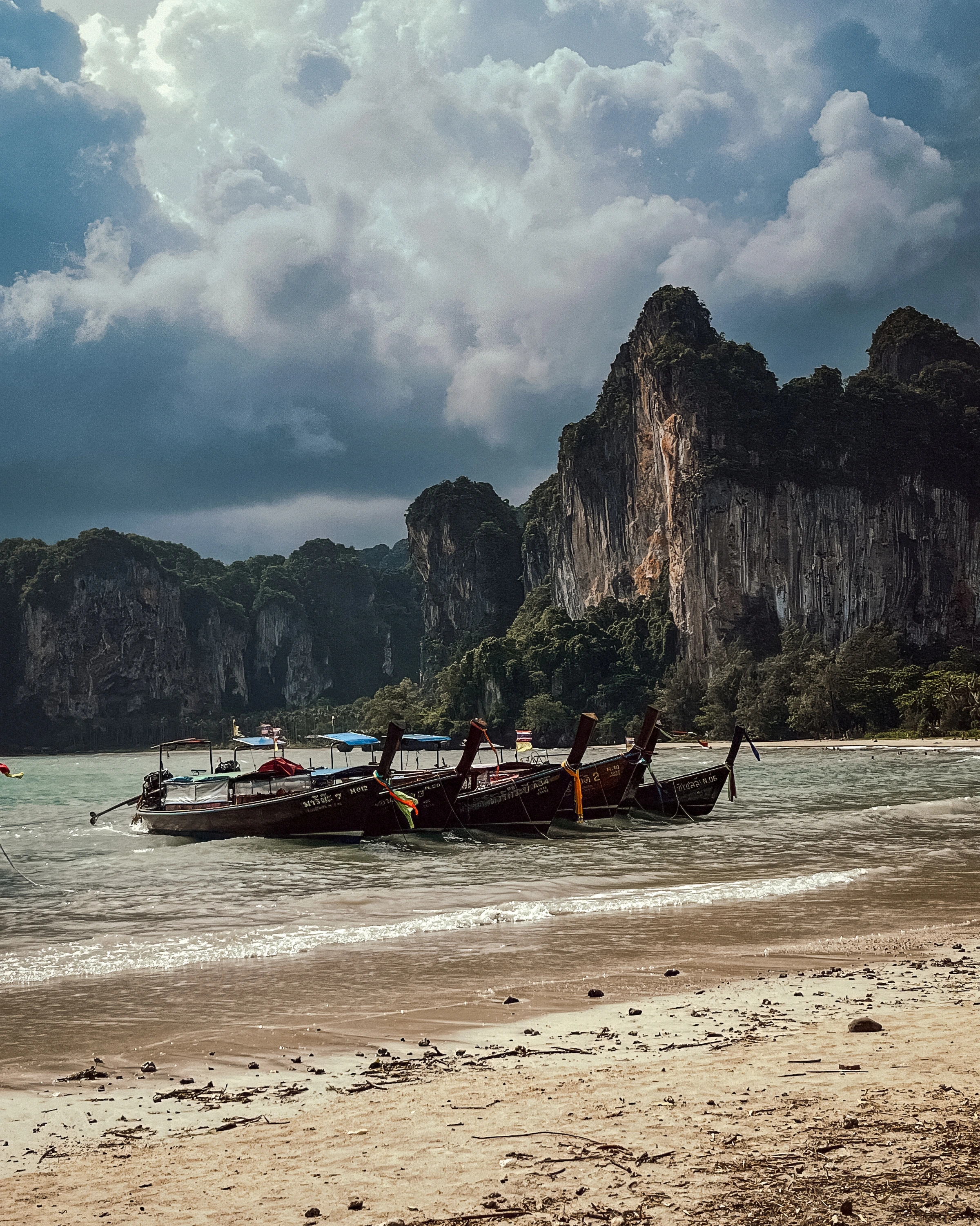Long wooden boats sit near a pale beach below tall vegetated cliffs and a cloudy sky.