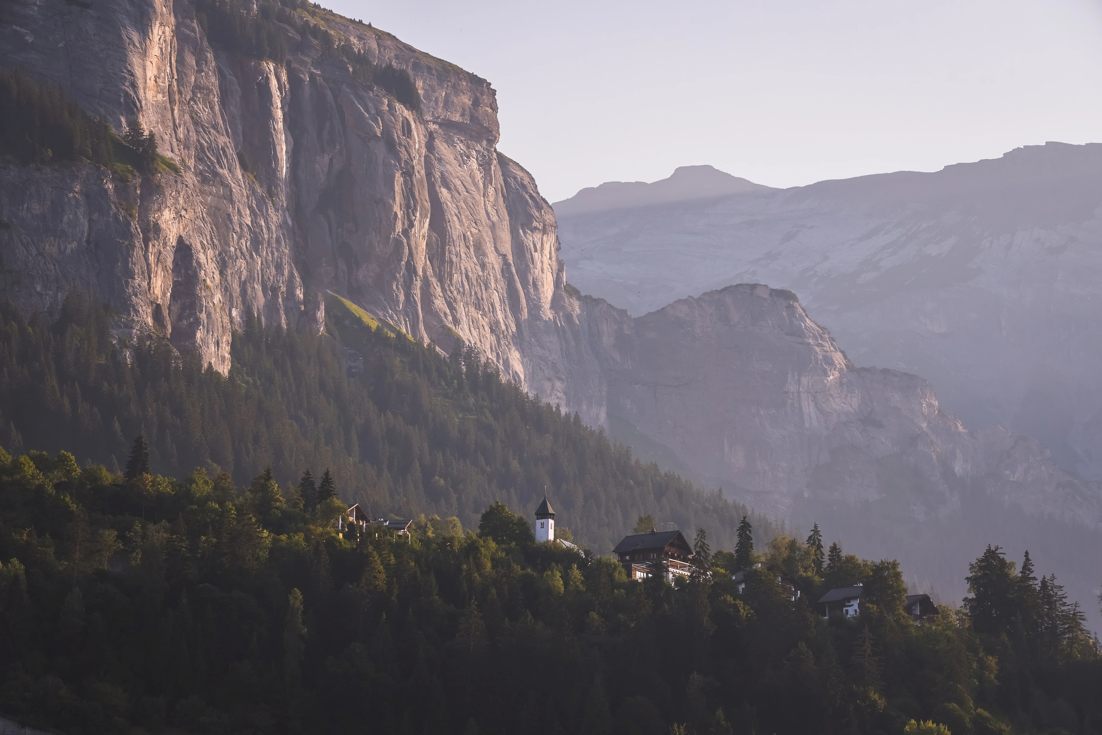 A pale church tower and small buildings sit among trees beneath a large sunlit cliff and hazy mountain layers.