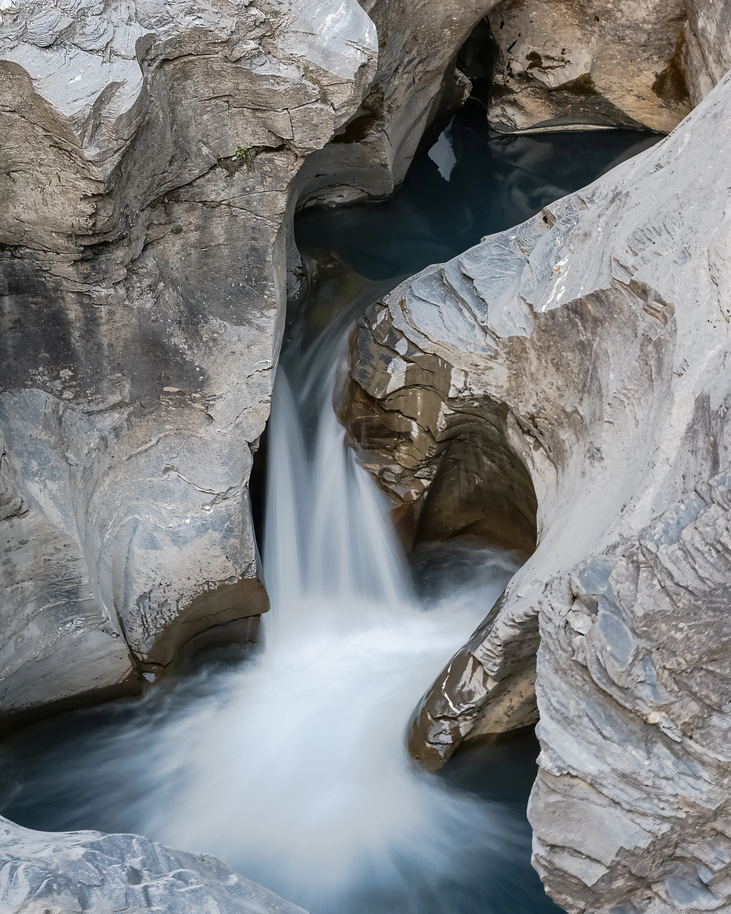 A narrow ribbon of water passes between curving grey rock surfaces and a dark upper pool.