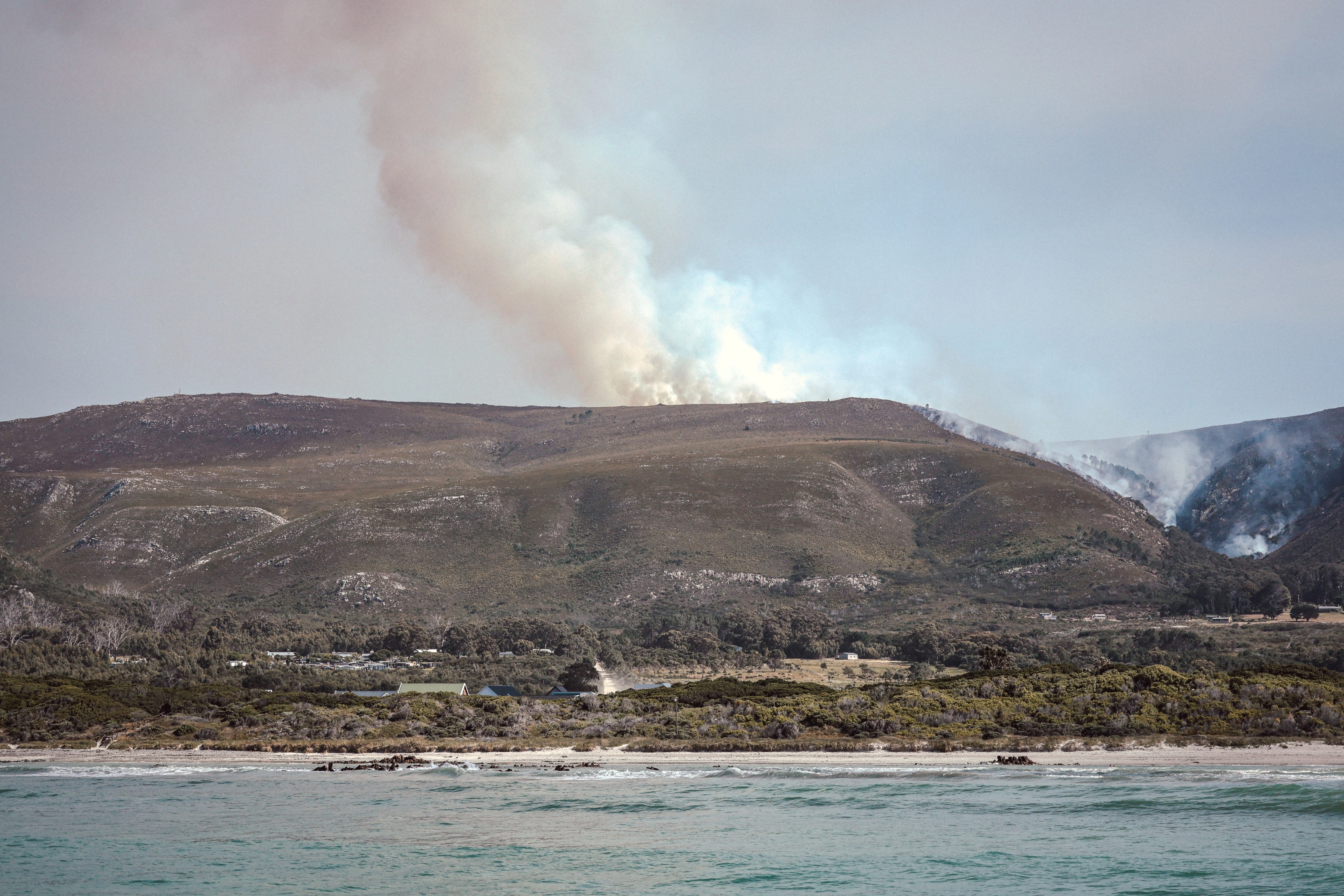 Buildings and a beach sit below a ridge, with a plume of smoke rising beyond the settlement.