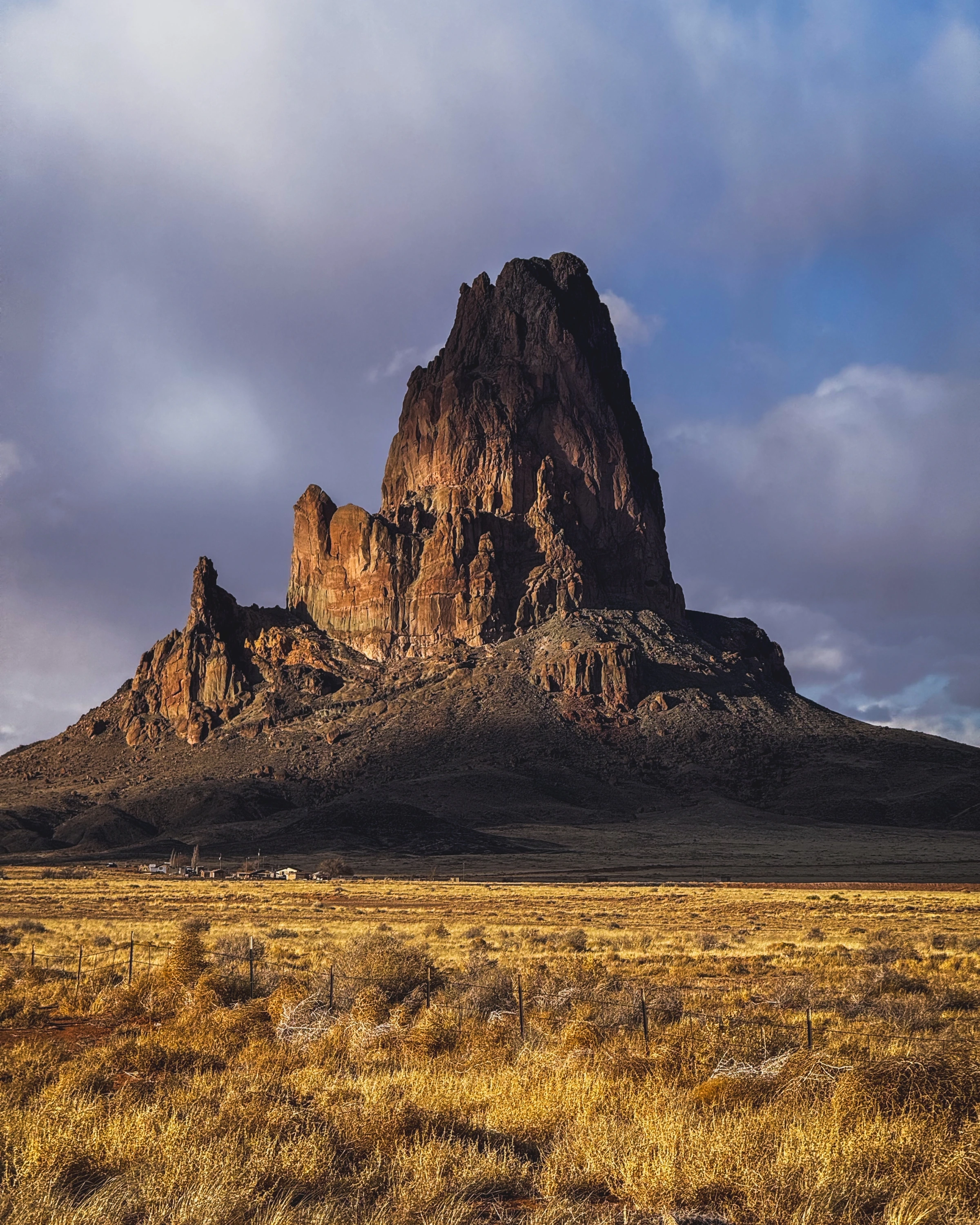 A tall banded rock spire rises above small buildings and grass, beneath a blue and cloudy sky.
