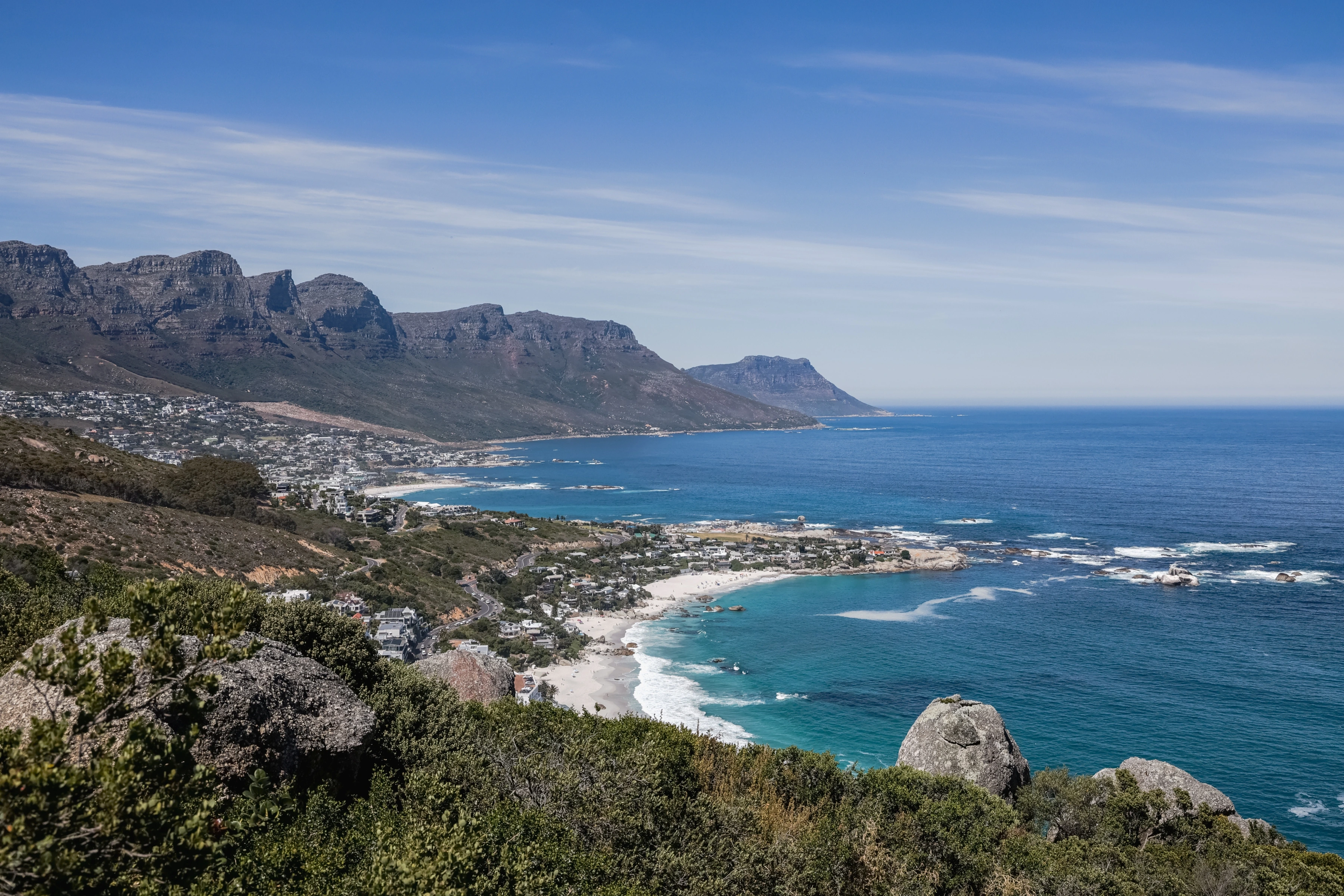 Foreground rocks overlook a long sweep of beach, coastal settlement, mountain ridge and blue ocean.