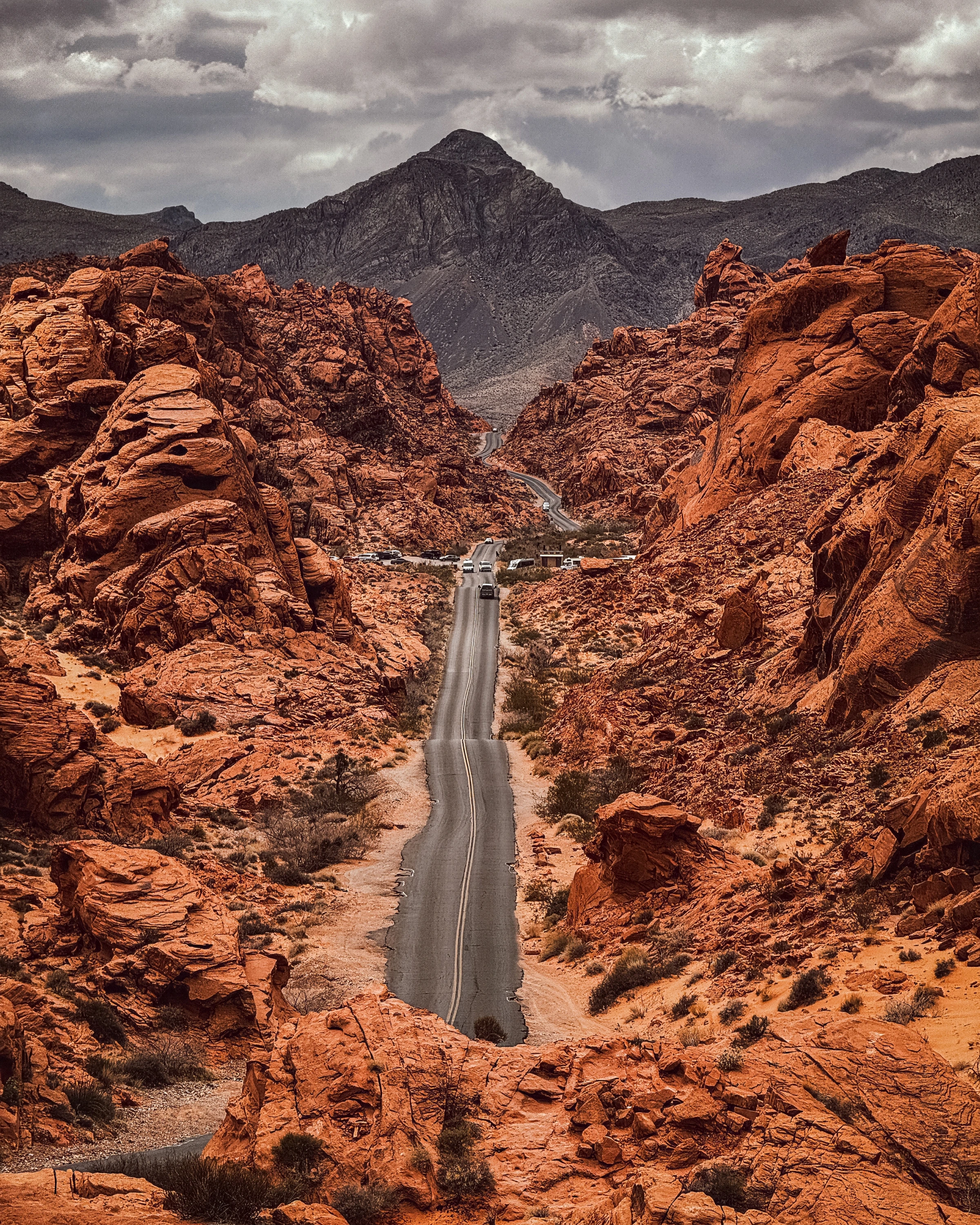 Vehicles follow a dipping road between orange rock walls beneath a grey mountain sky.