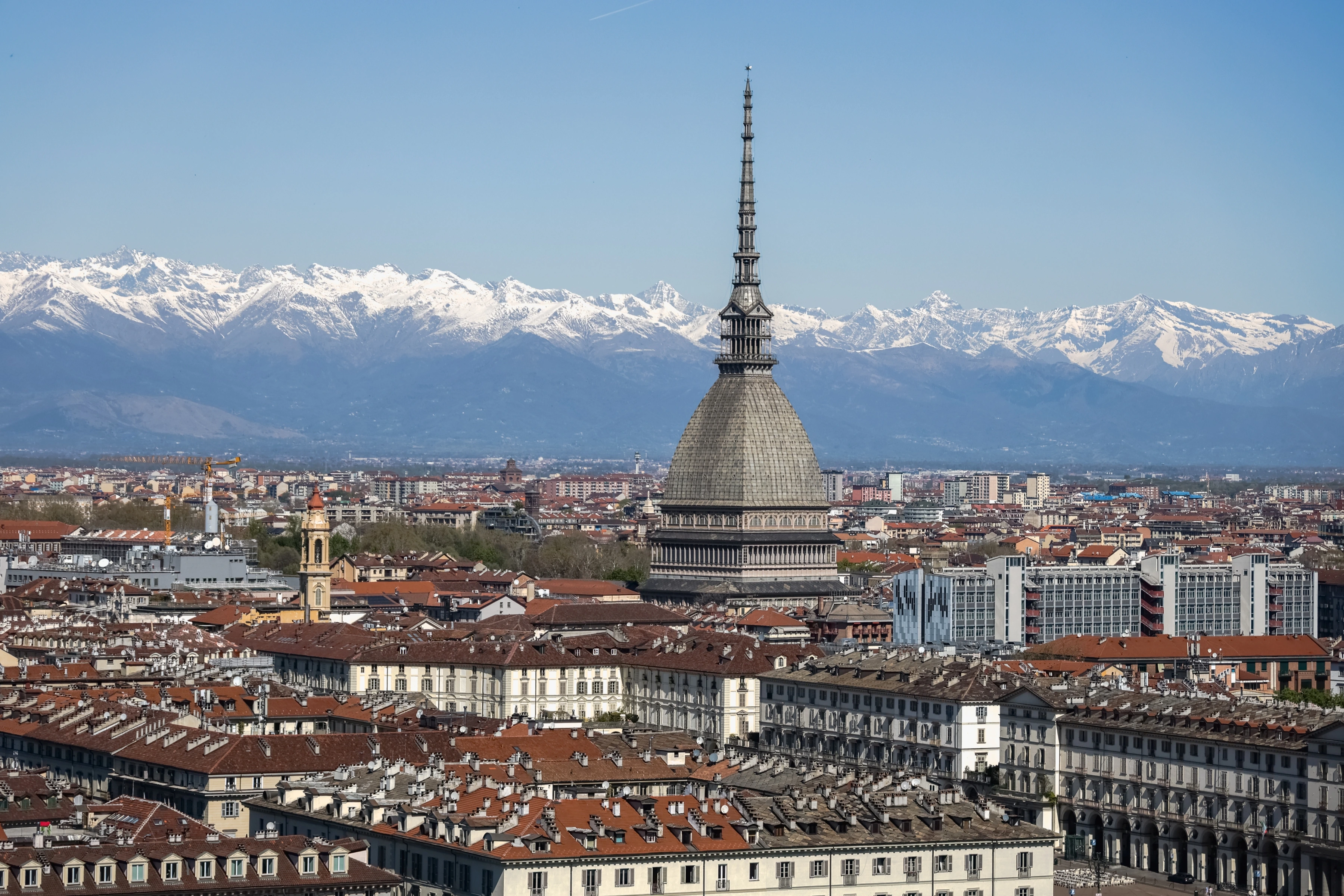 A tall pointed spire rises from dense city roofs beneath a broad line of snowy mountains.