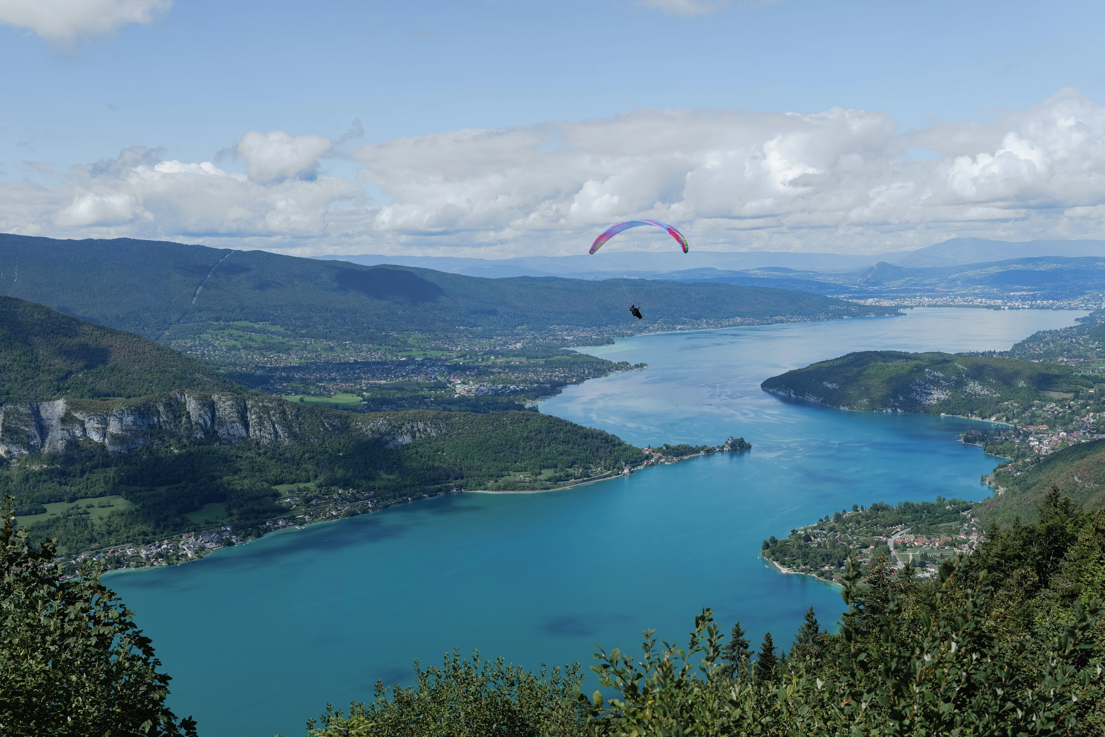 A paraglider crosses above a broad lake enclosed by hills and settlements.