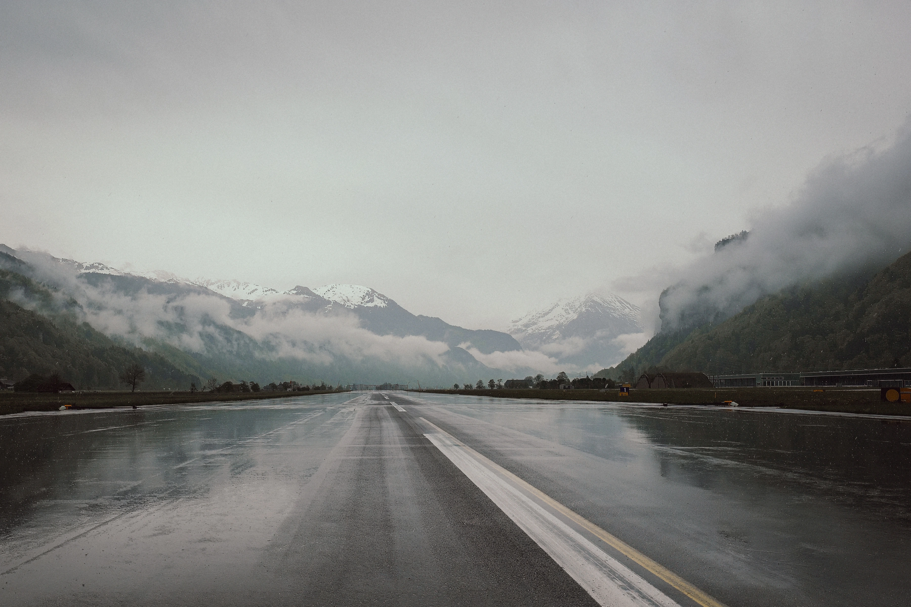 Runway markings recede across wet pavement between mountains partly hidden by cloud.