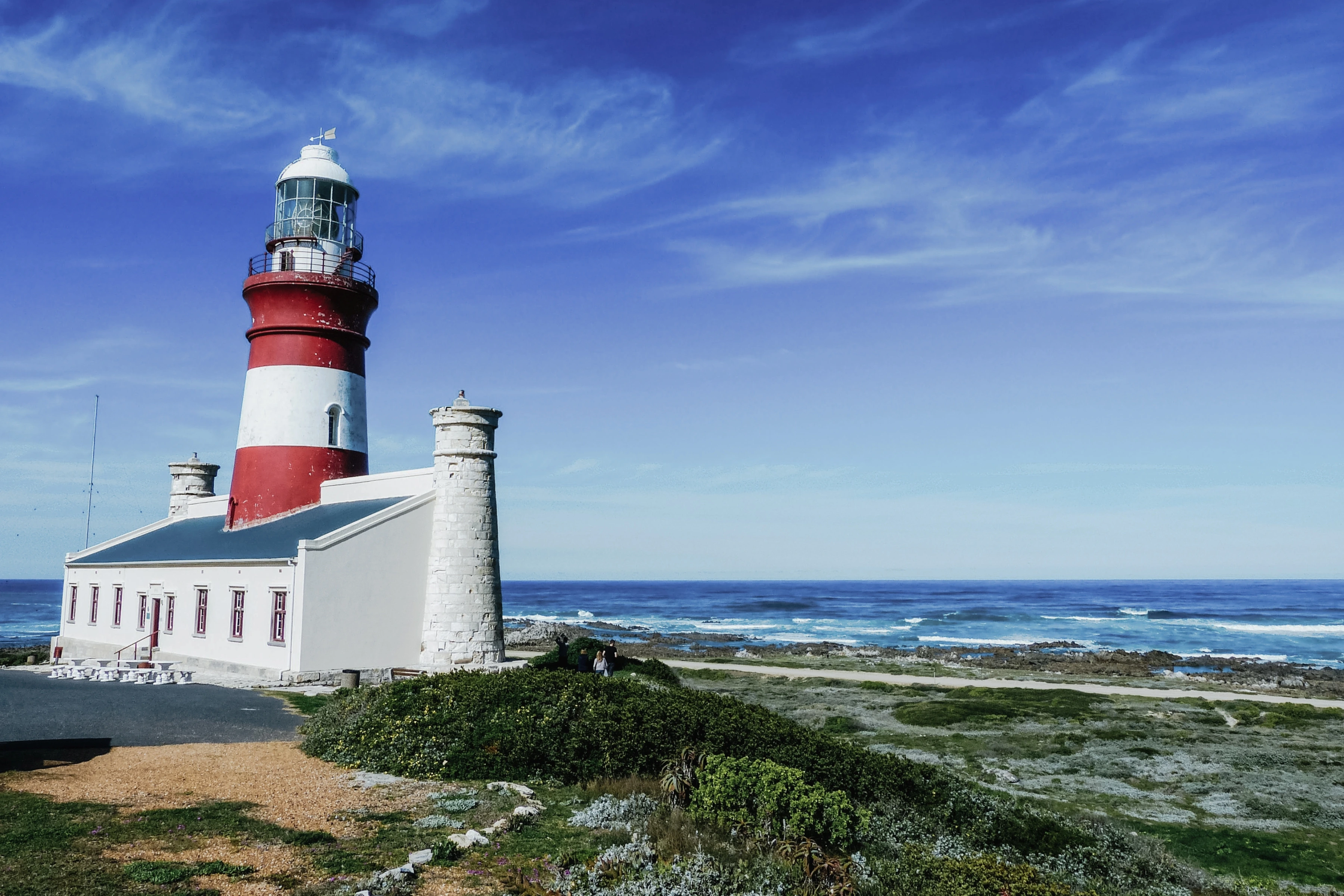 A red-and-white lighthouse and low building stand beside rough sea and pale ground vegetation.