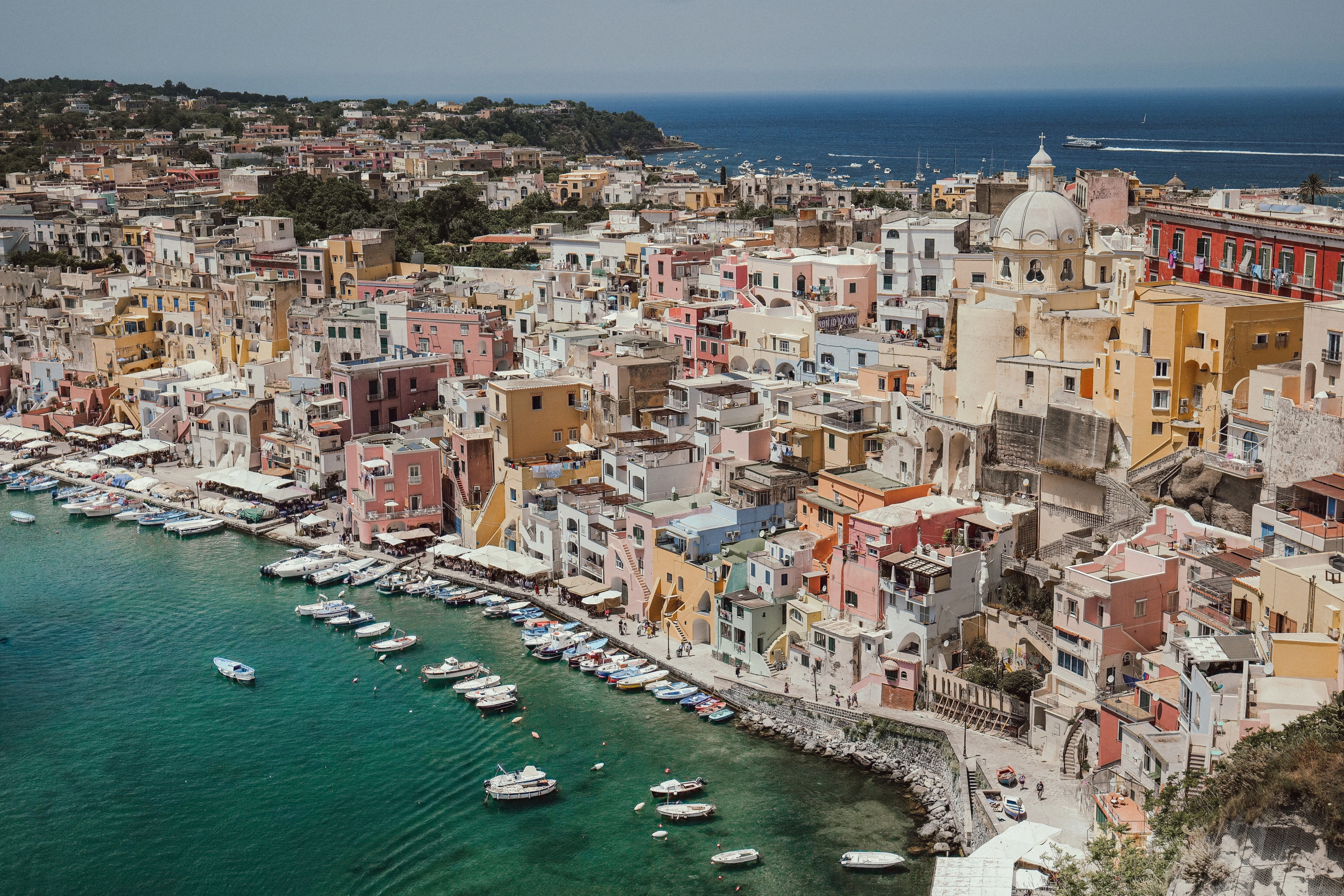 Small boats lie below closely packed coloured houses and a domed building by the sea.