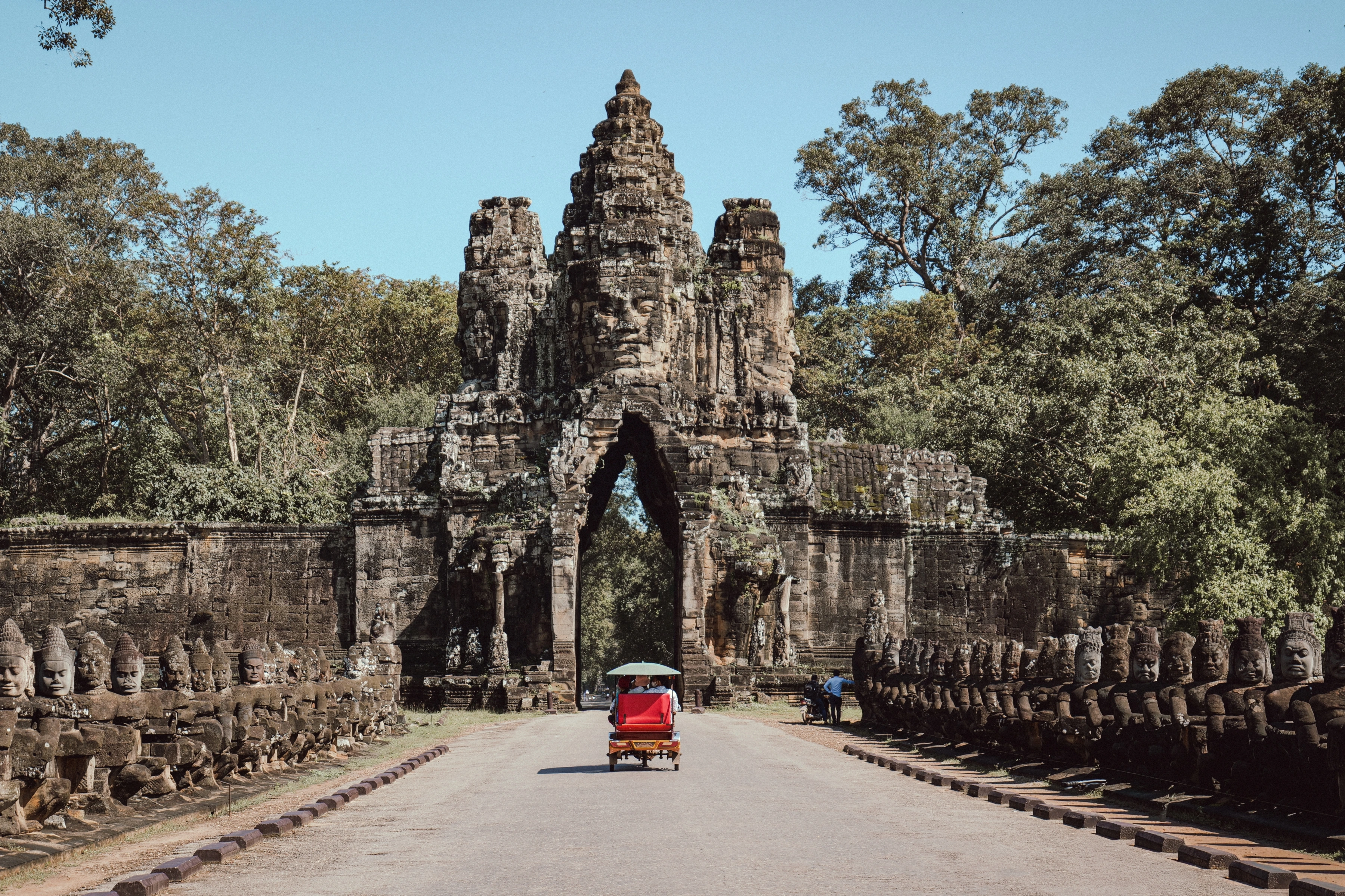 A red three-wheeled vehicle passes through a stone gateway between rows of statues.