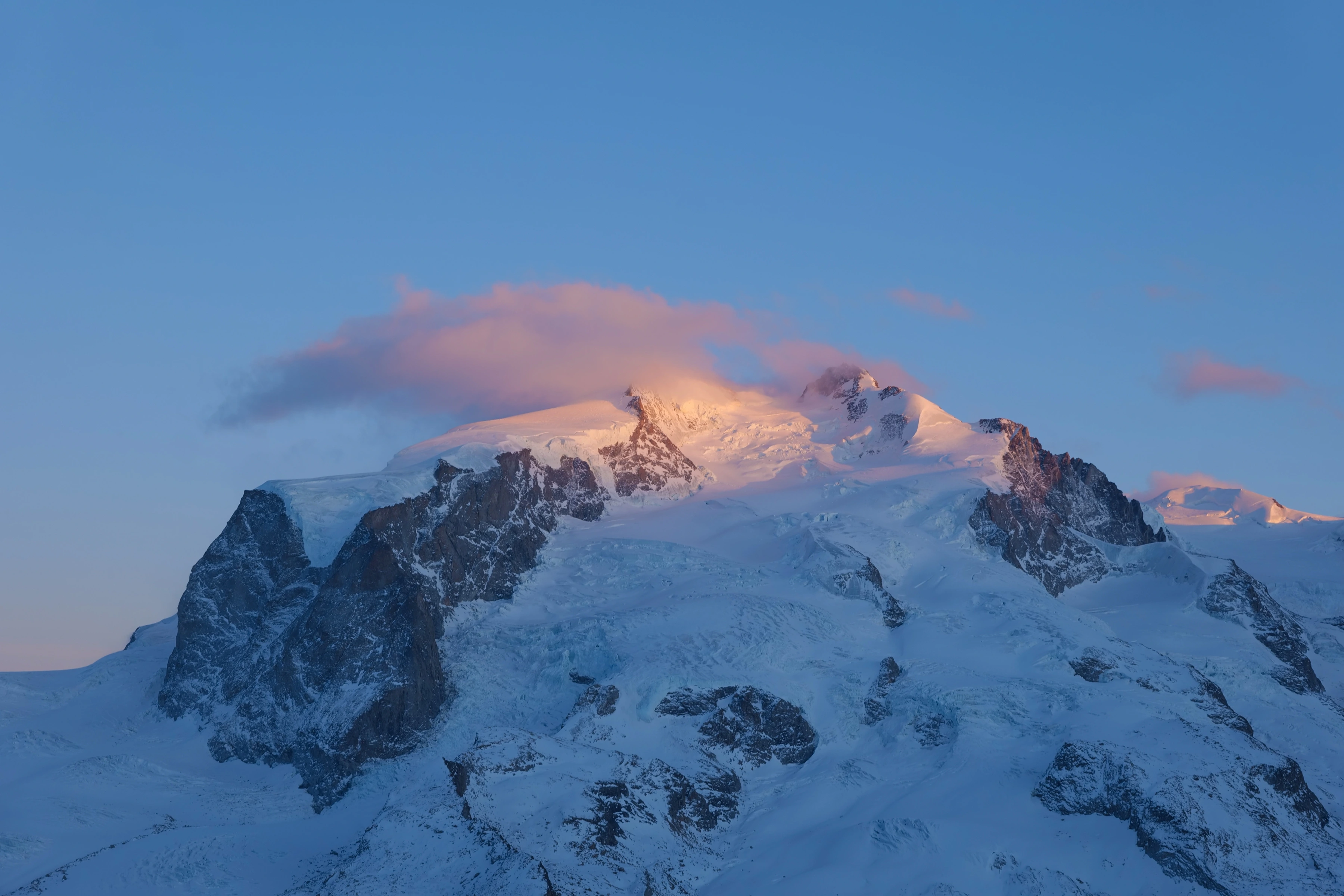 Warm light touches a snow-covered mountain crest above cooler blue snow and shadow.