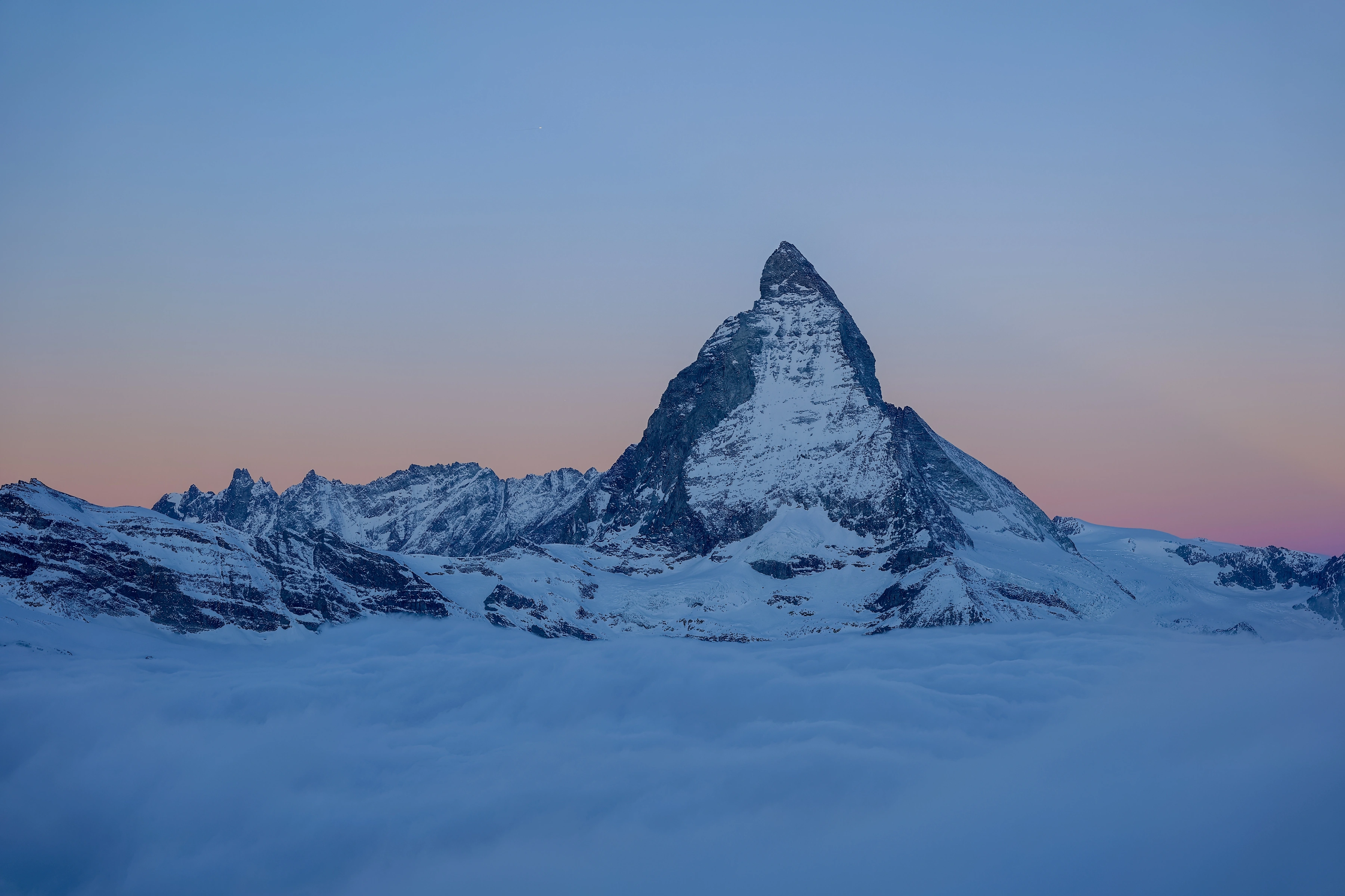A triangular snow-covered peak rises above a continuous cloud layer under a pale warm-to-blue sky.