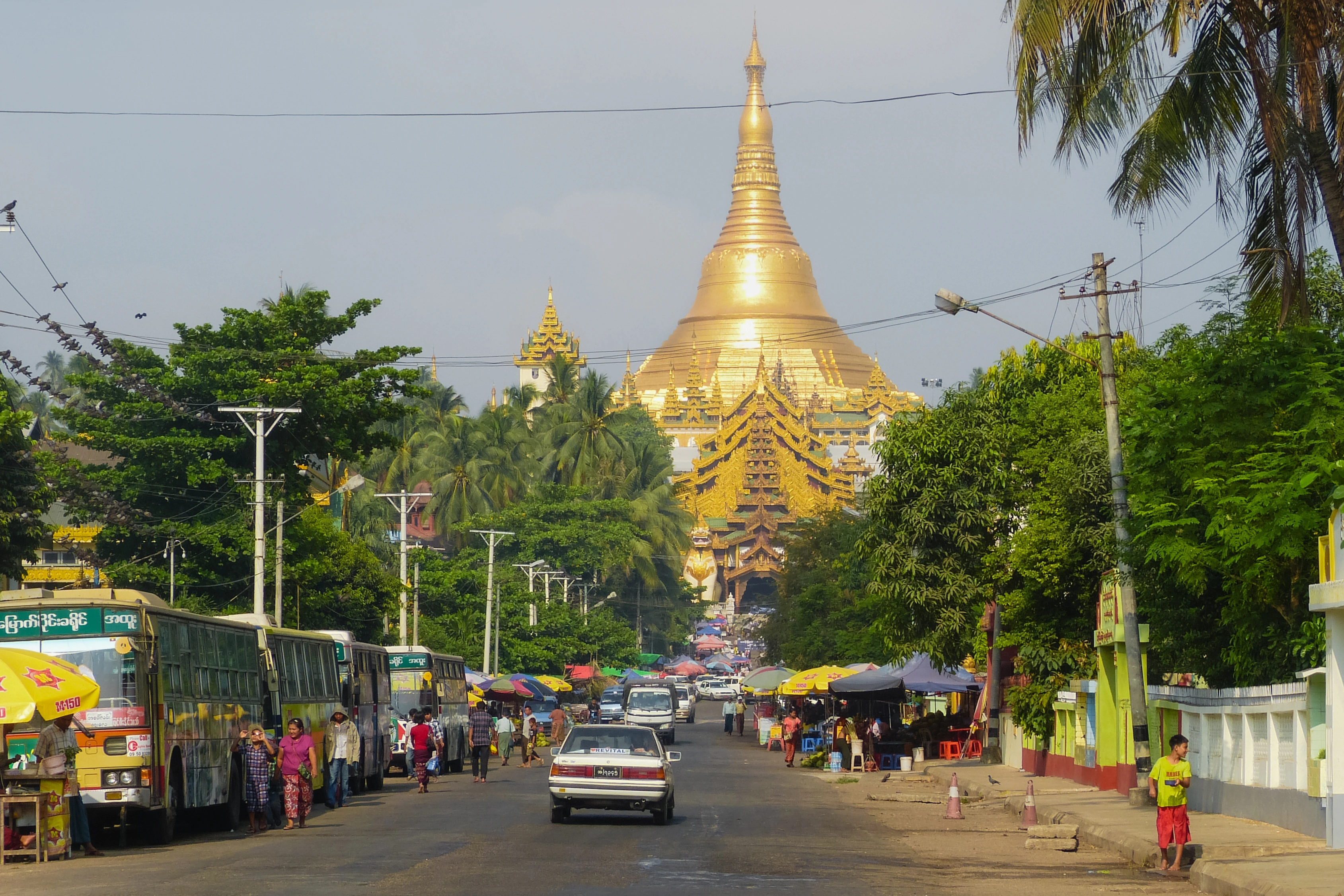 Buses, cars, market umbrellas and people occupy a street beneath a large golden tower.