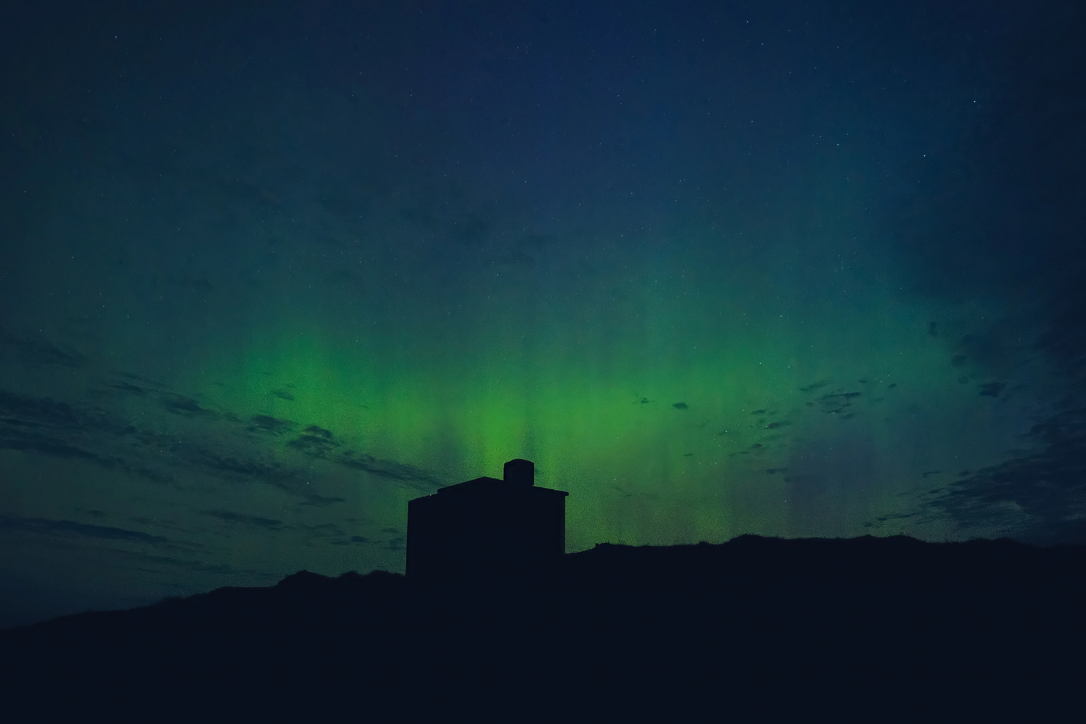 A green aurora stretches over a small hut and dark ground.