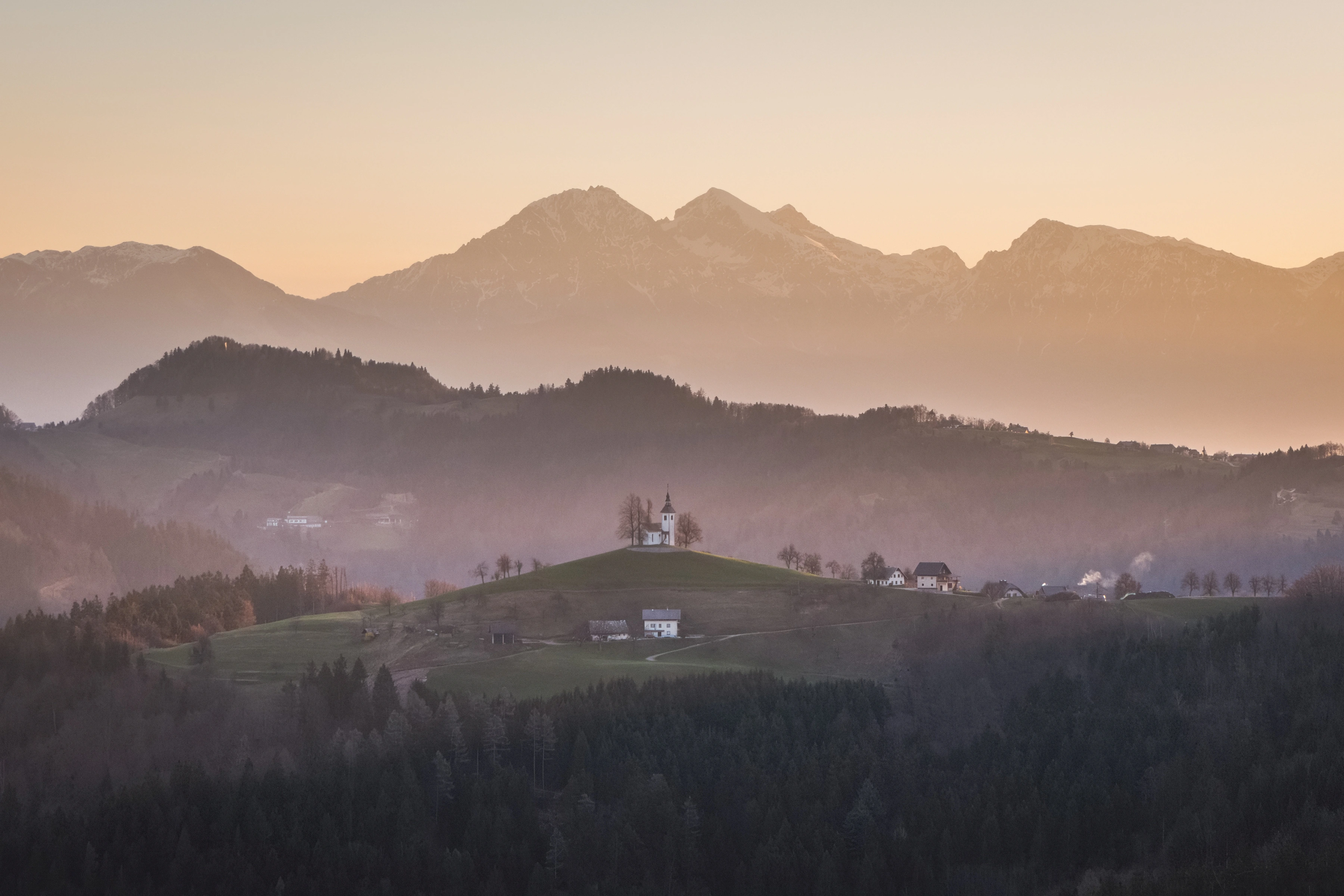 A small church stands on a hill amid layered woods, houses and hazy mountain ridges.