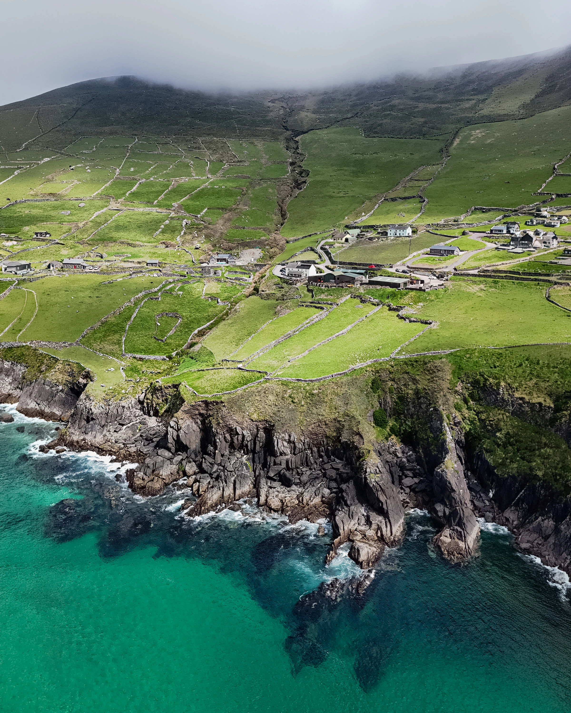 Stone-walled fields and houses sit above dark cliffs and turquoise water under low cloud.