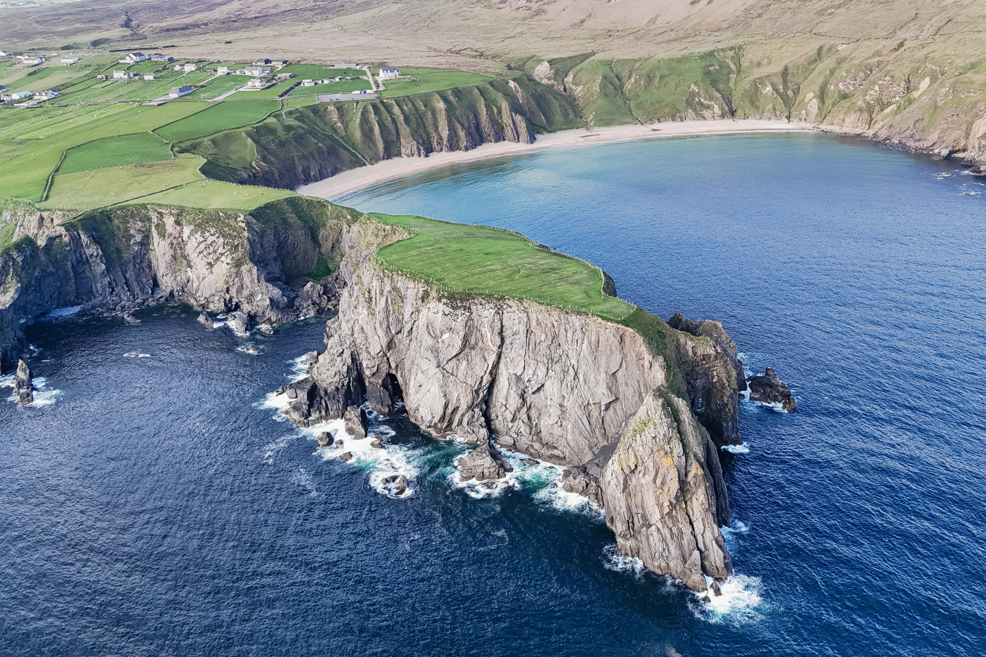 A green headland with rock arches projects into the sea beside a curving bay.