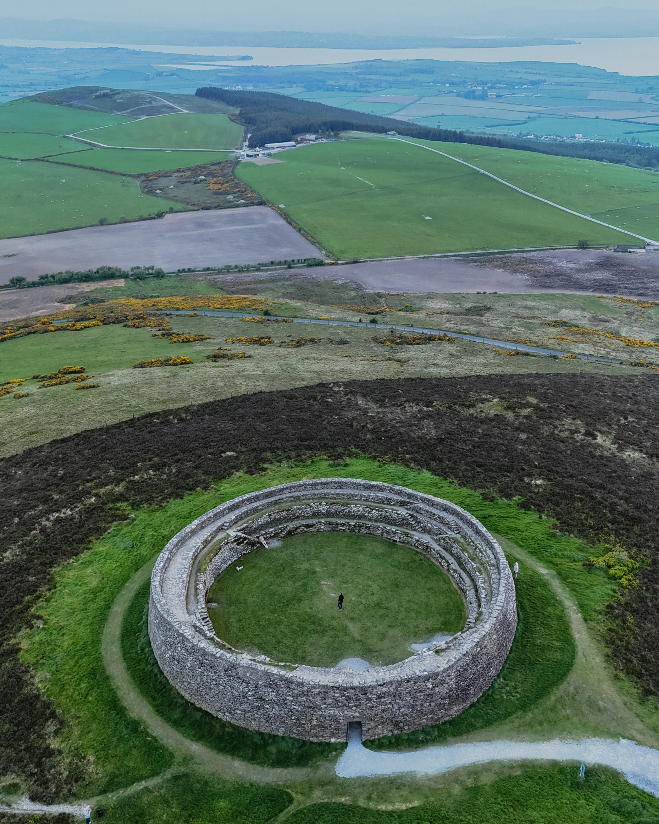 A small person stands inside a circular stone enclosure above a patchwork of fields, roads and distant water.