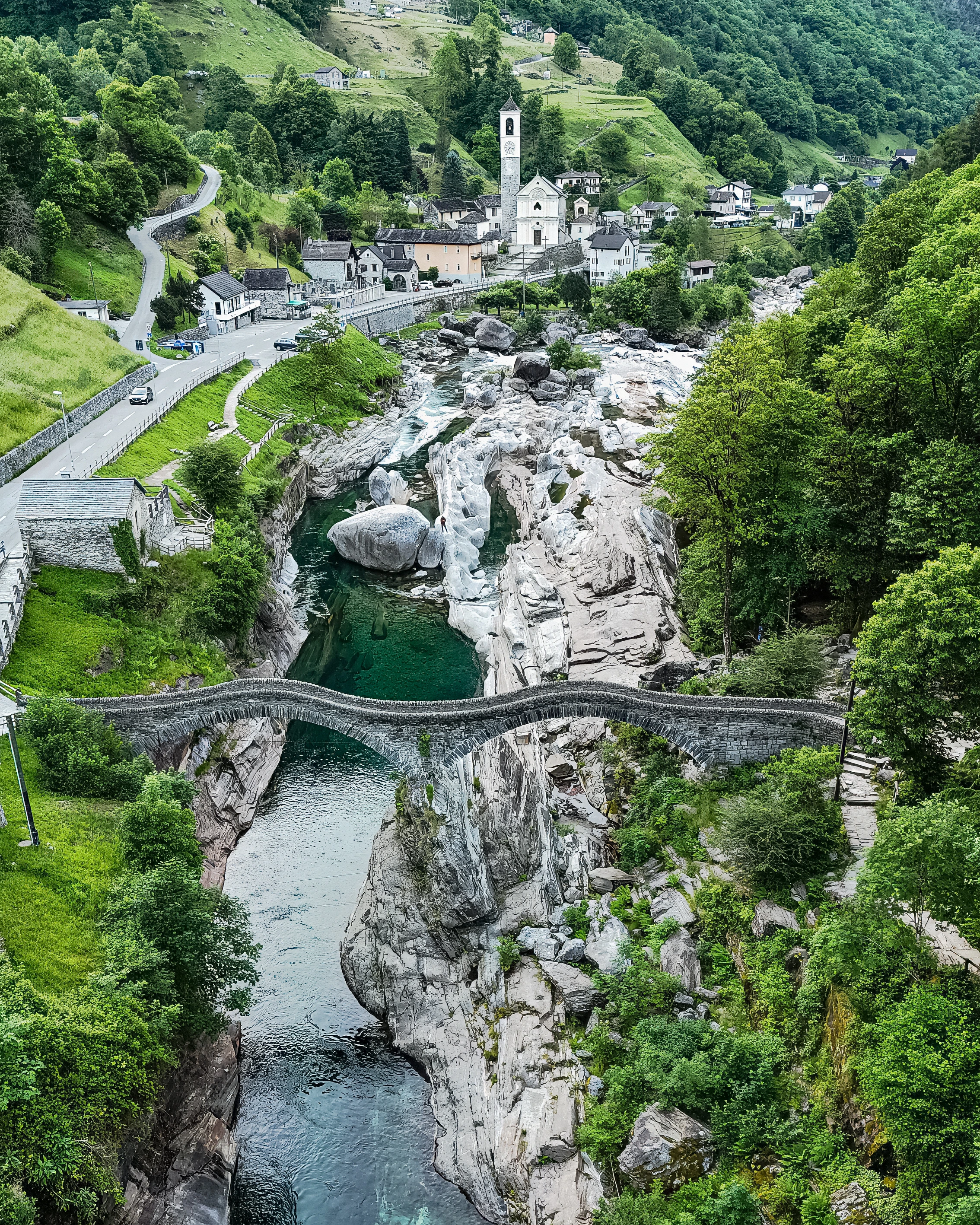 An arched bridge crosses a river beside exposed pale rock, small people and a village church.