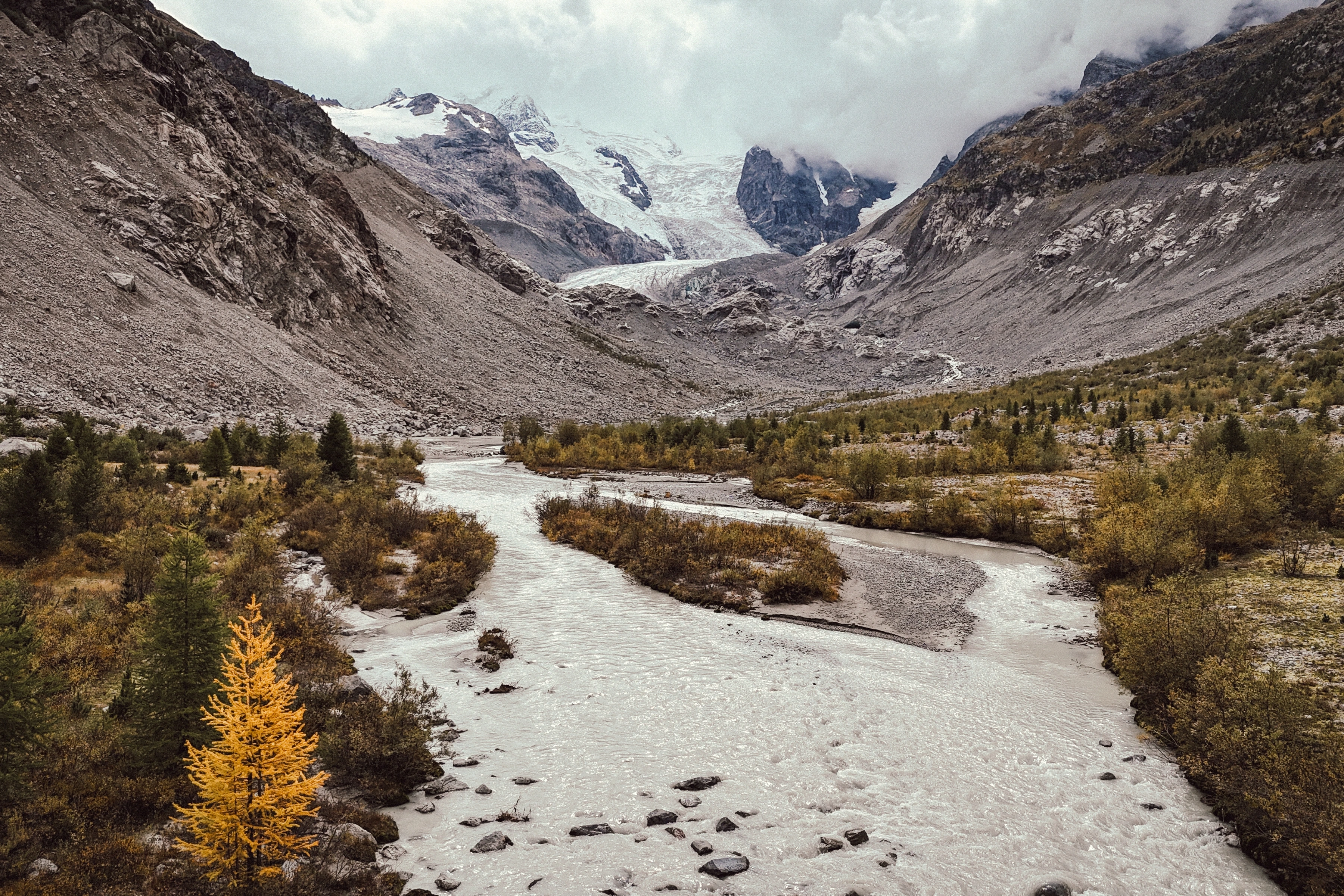 A pale braided river curves through a gravel valley toward a glacier between steep mountain slopes.