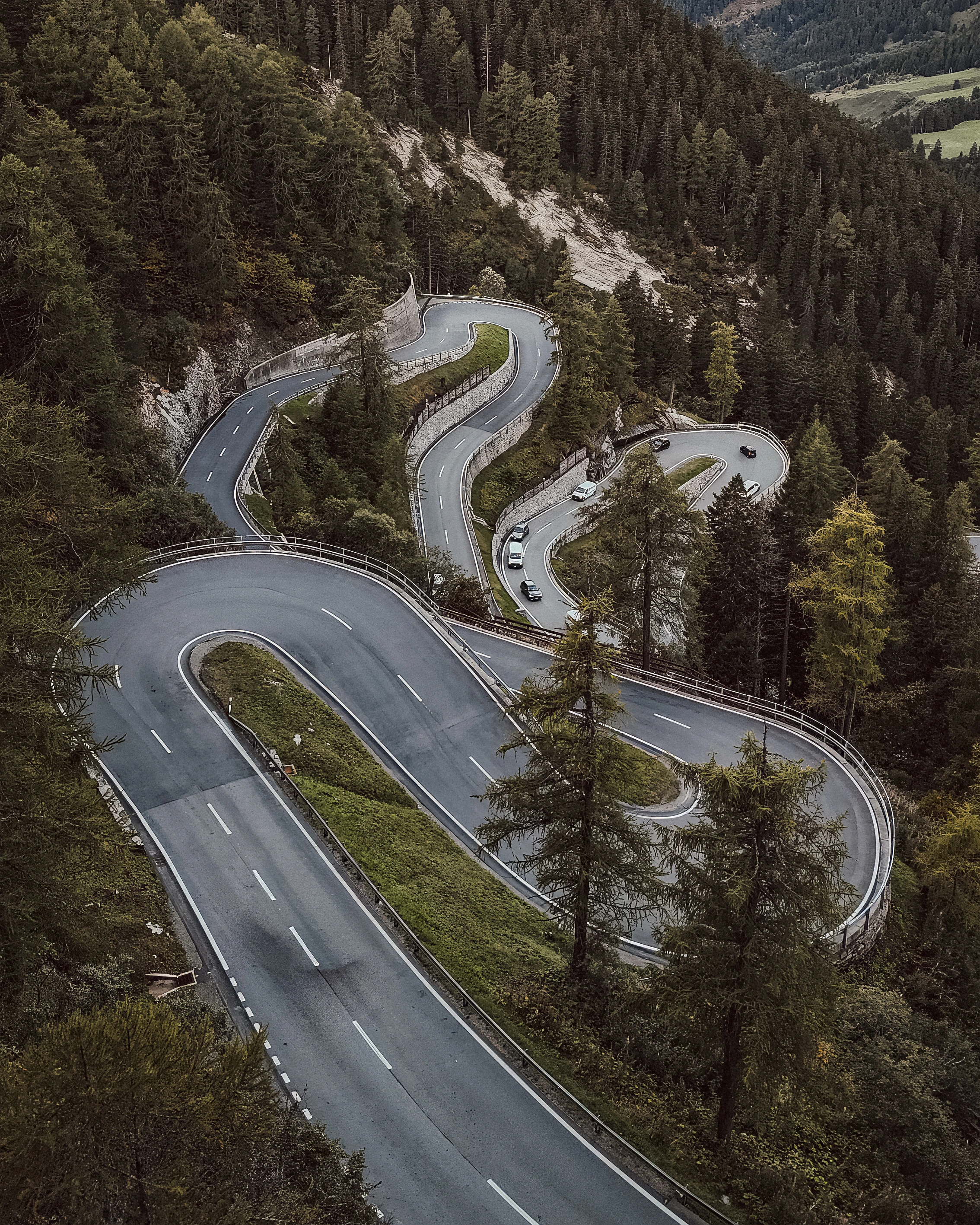 A road makes repeated hairpin bends through dense forest with small cars for scale.