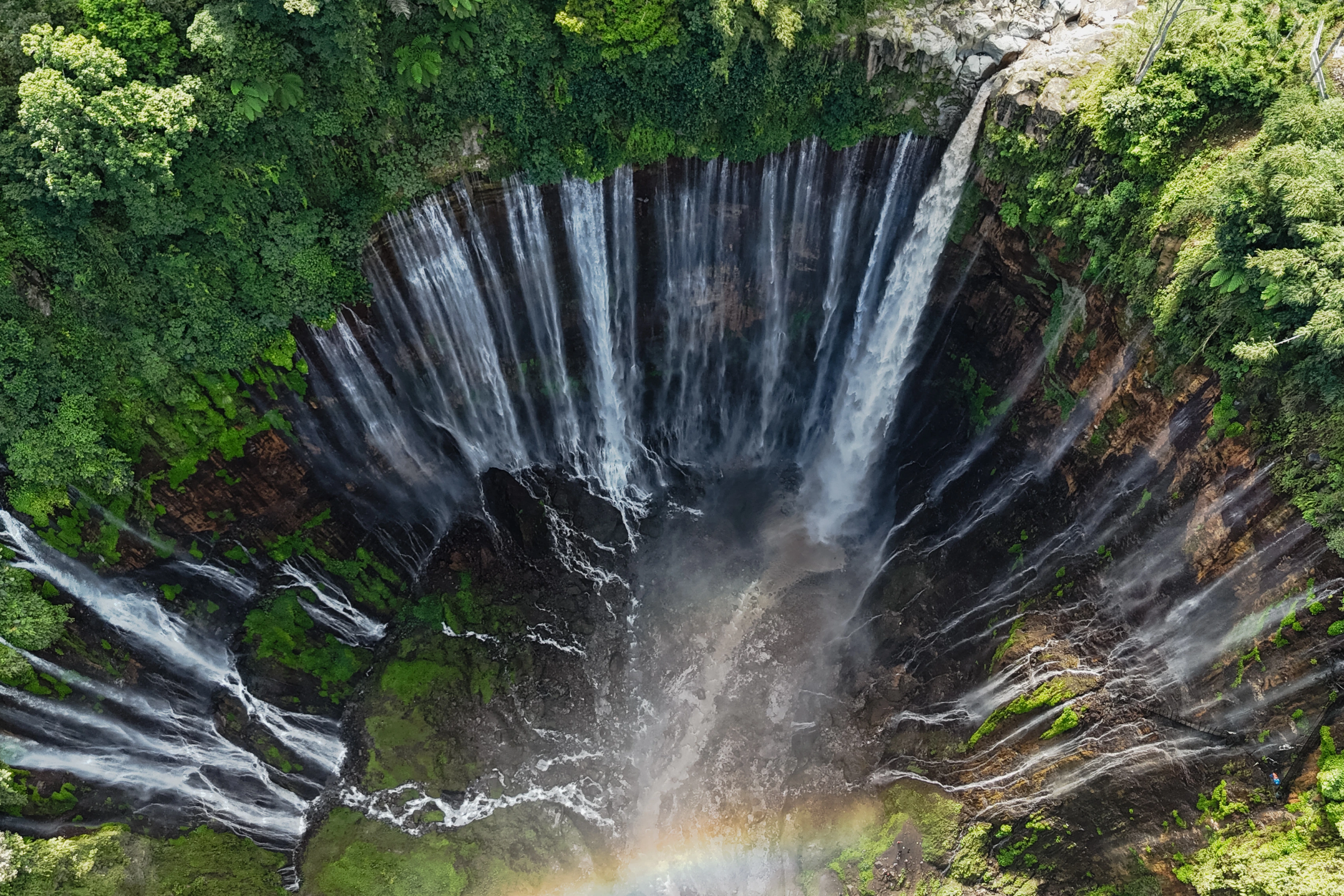 An overhead view shows many streams dropping into a green-sided basin with a faint rainbow below.